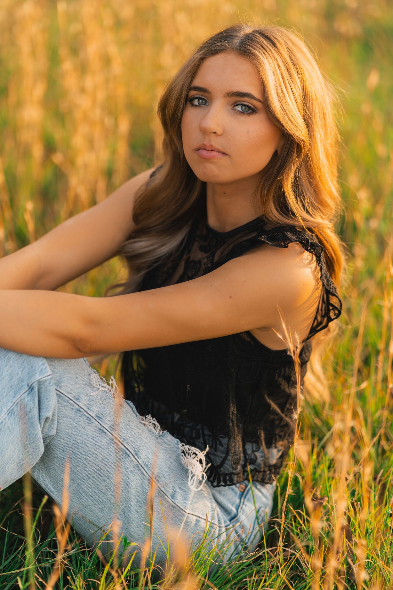 Portrait of someone in a black top and distressed jeans sitting in a golden sunlit field at sunset.