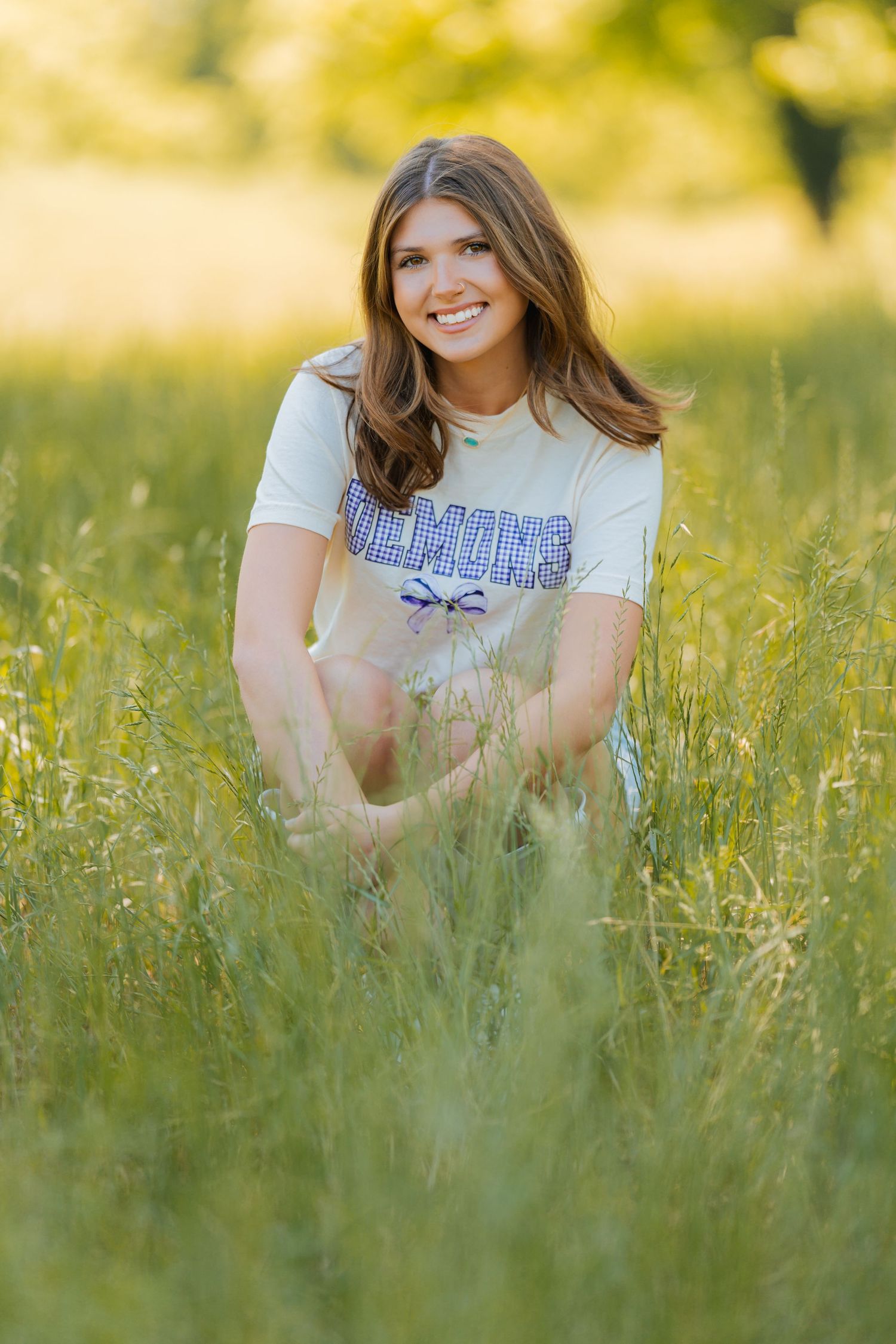 A person in a University of Maine t-shirt sits smiling in tall grass during golden hour photography.