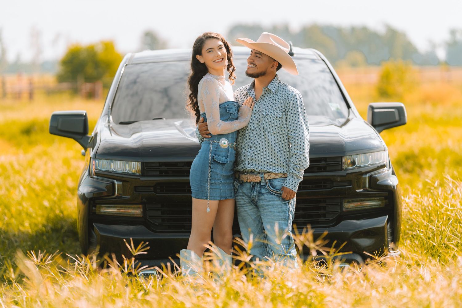 A couple embraces in front of a black car in a sunlit field of golden autumn grass.