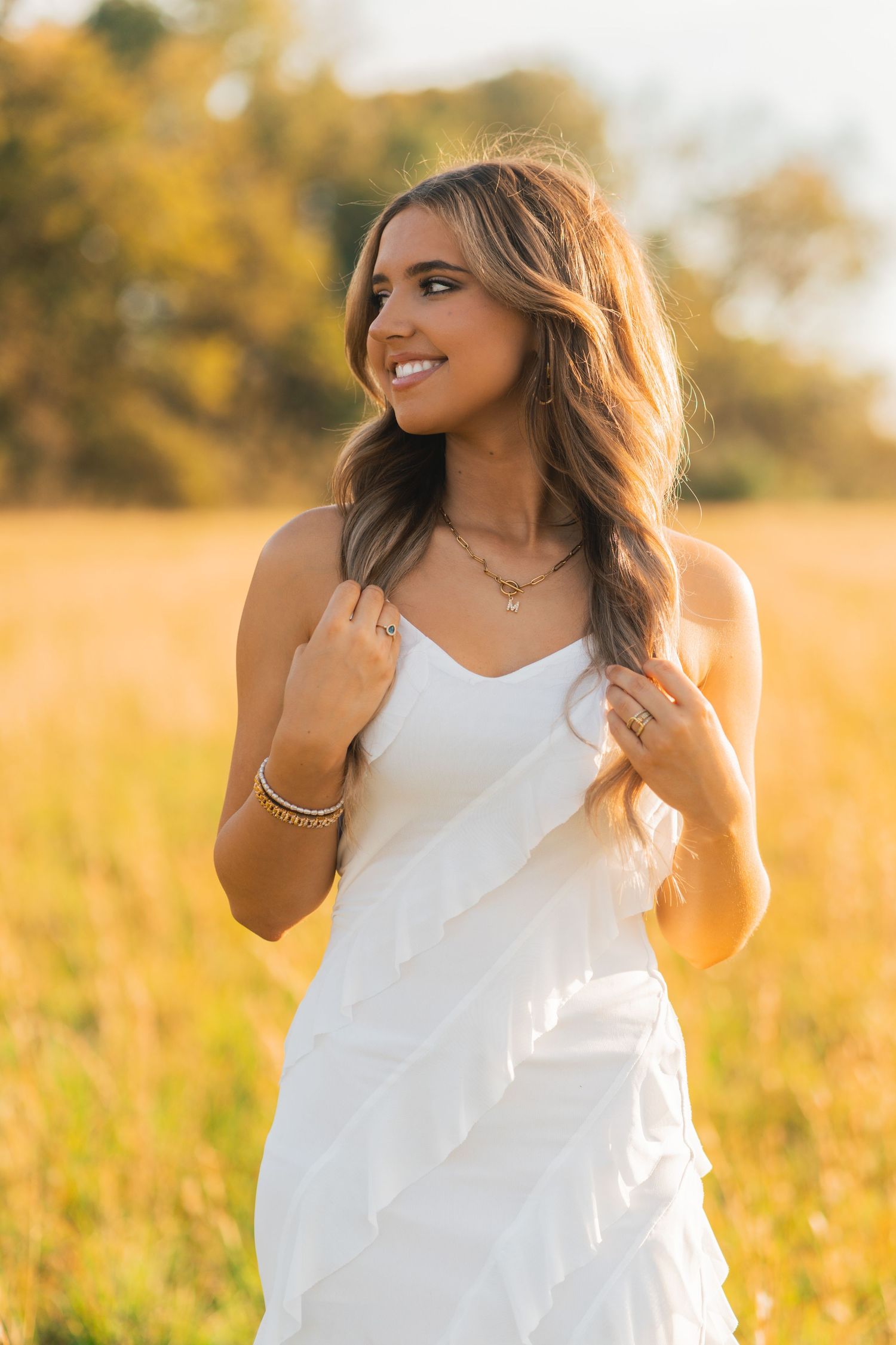 A woman in a white sundress poses in a golden field at sunset while smiling and touching her hair.