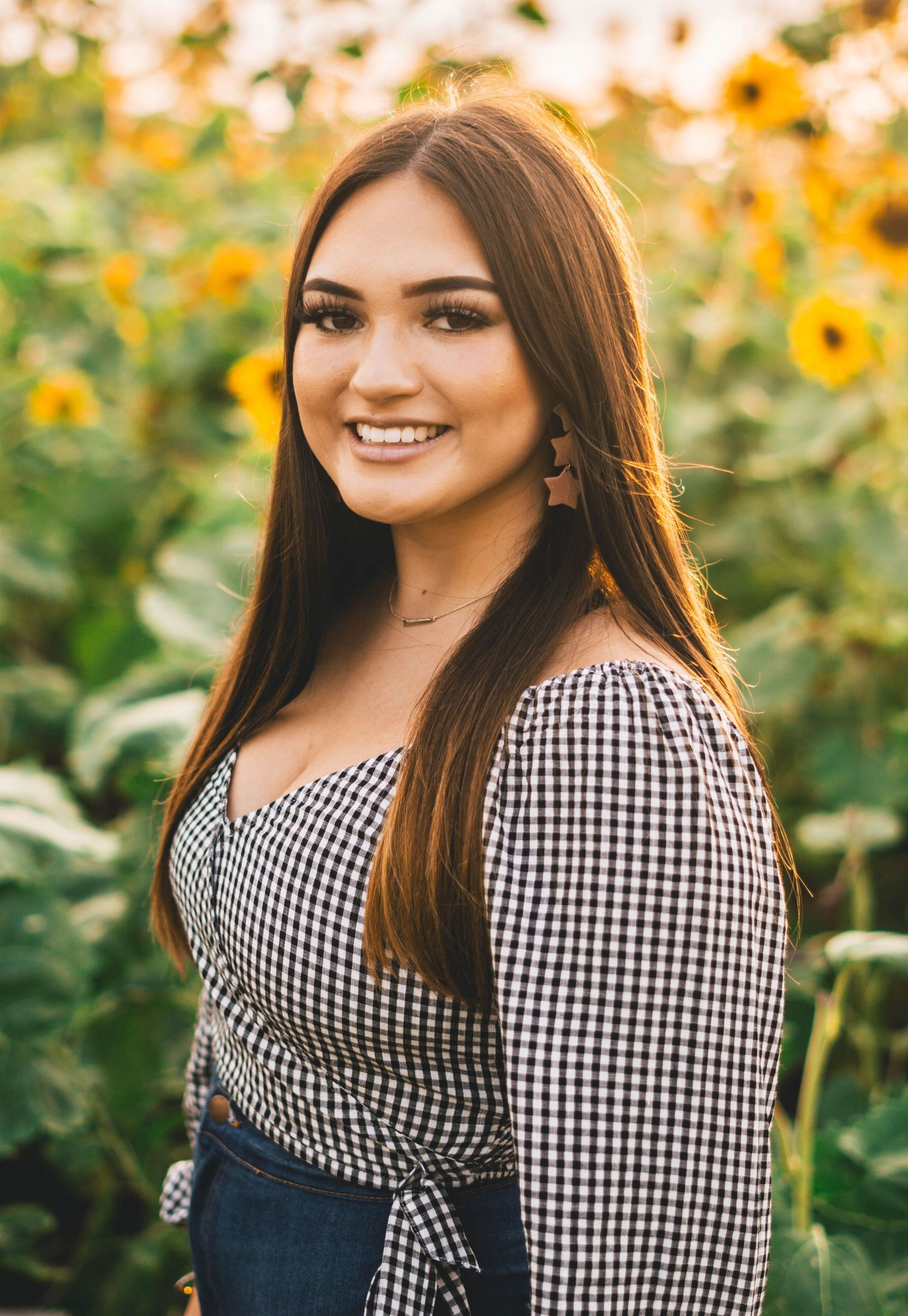 A smiling woman in a black and white gingham wrap top poses in front of blooming sunflowers during golden hour.