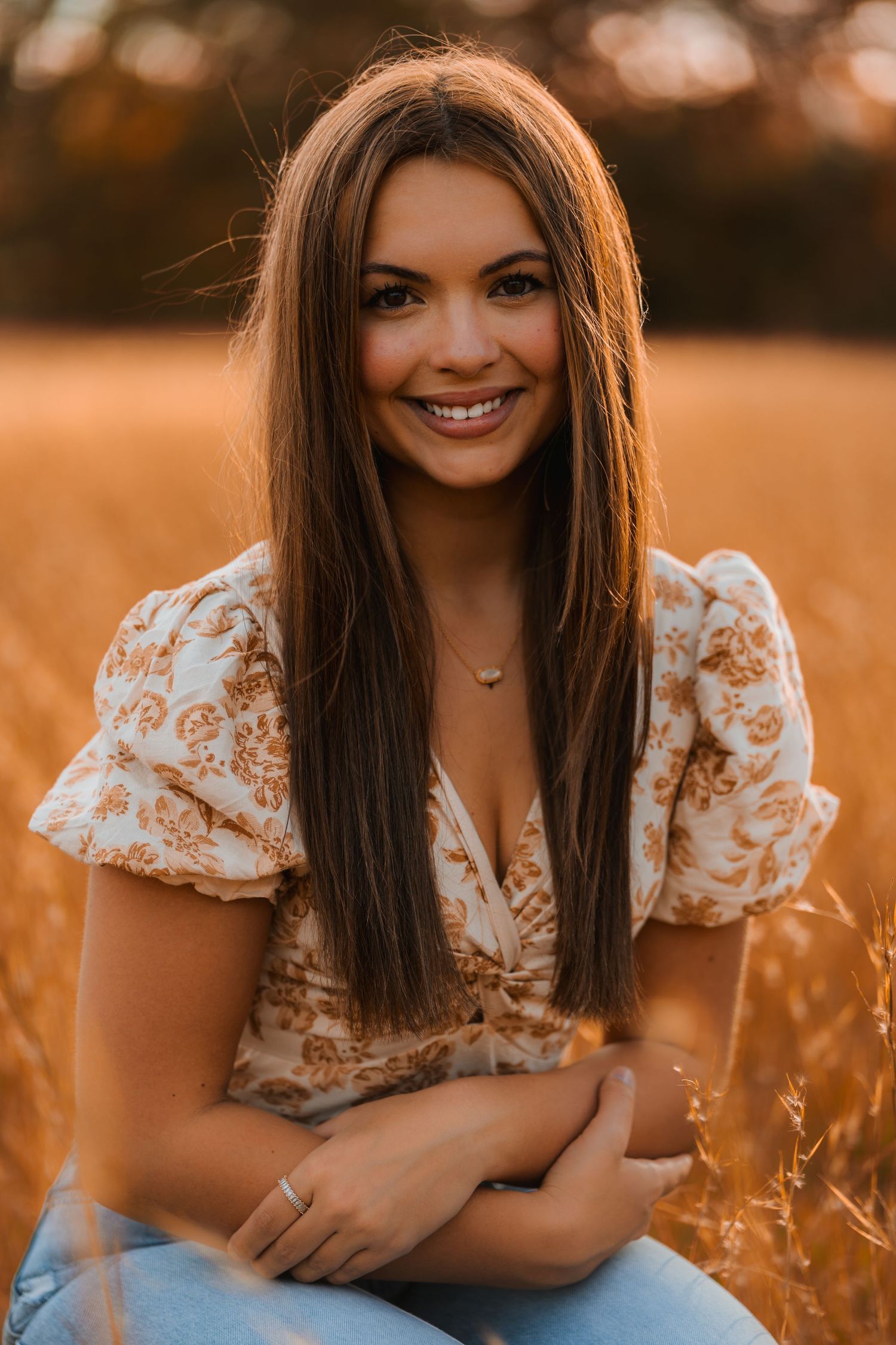 Portrait of a woman in a floral top sitting in a golden field at sunset with long brown hair and a delicate necklace.