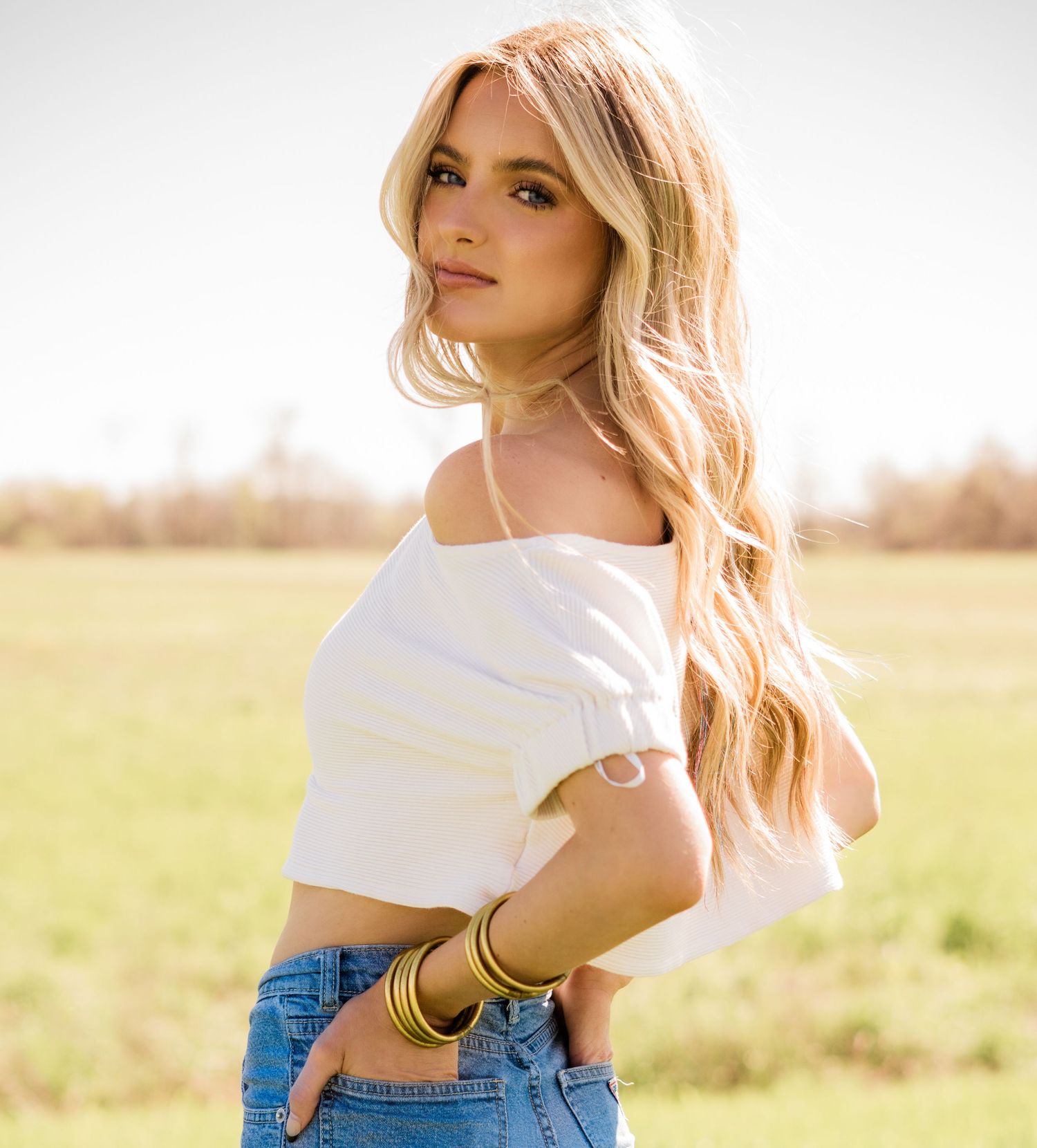 Person in cropped white off-shoulder top and jeans poses in sunny field with long blonde hair.