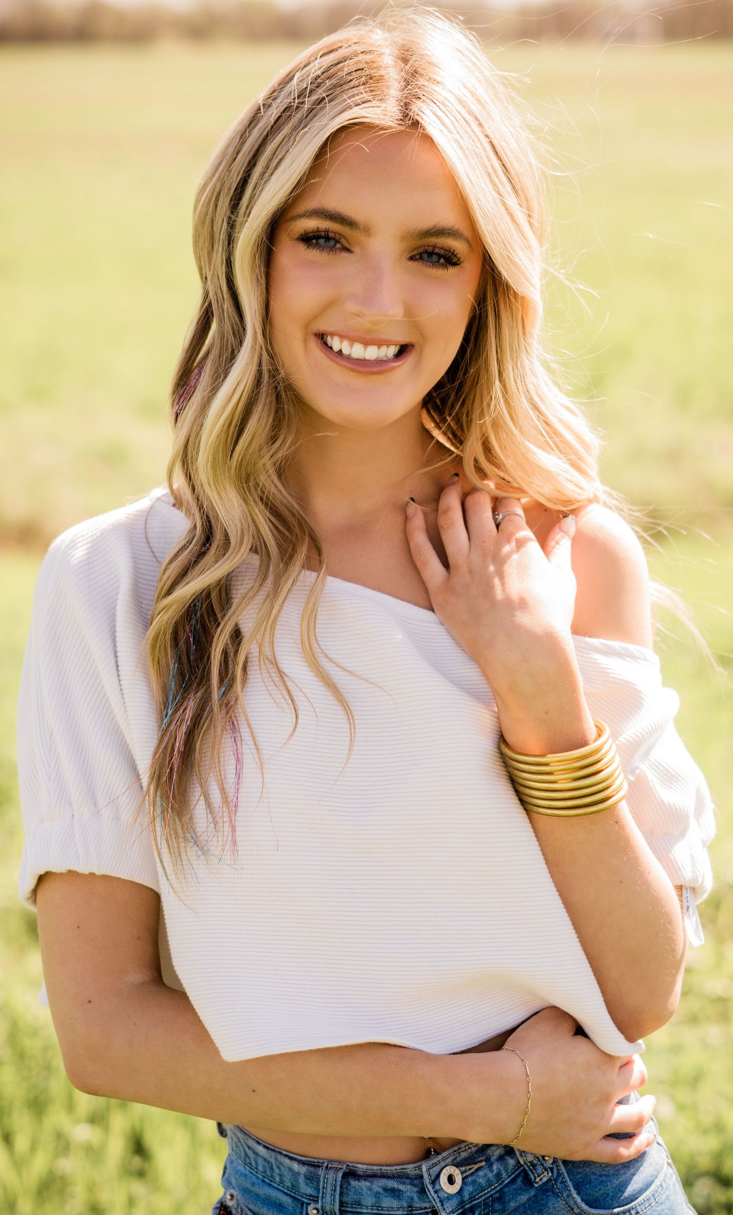 Portrait in warm sunlight showing blonde wavy hair, white top and gold bangles against a natural outdoor setting.