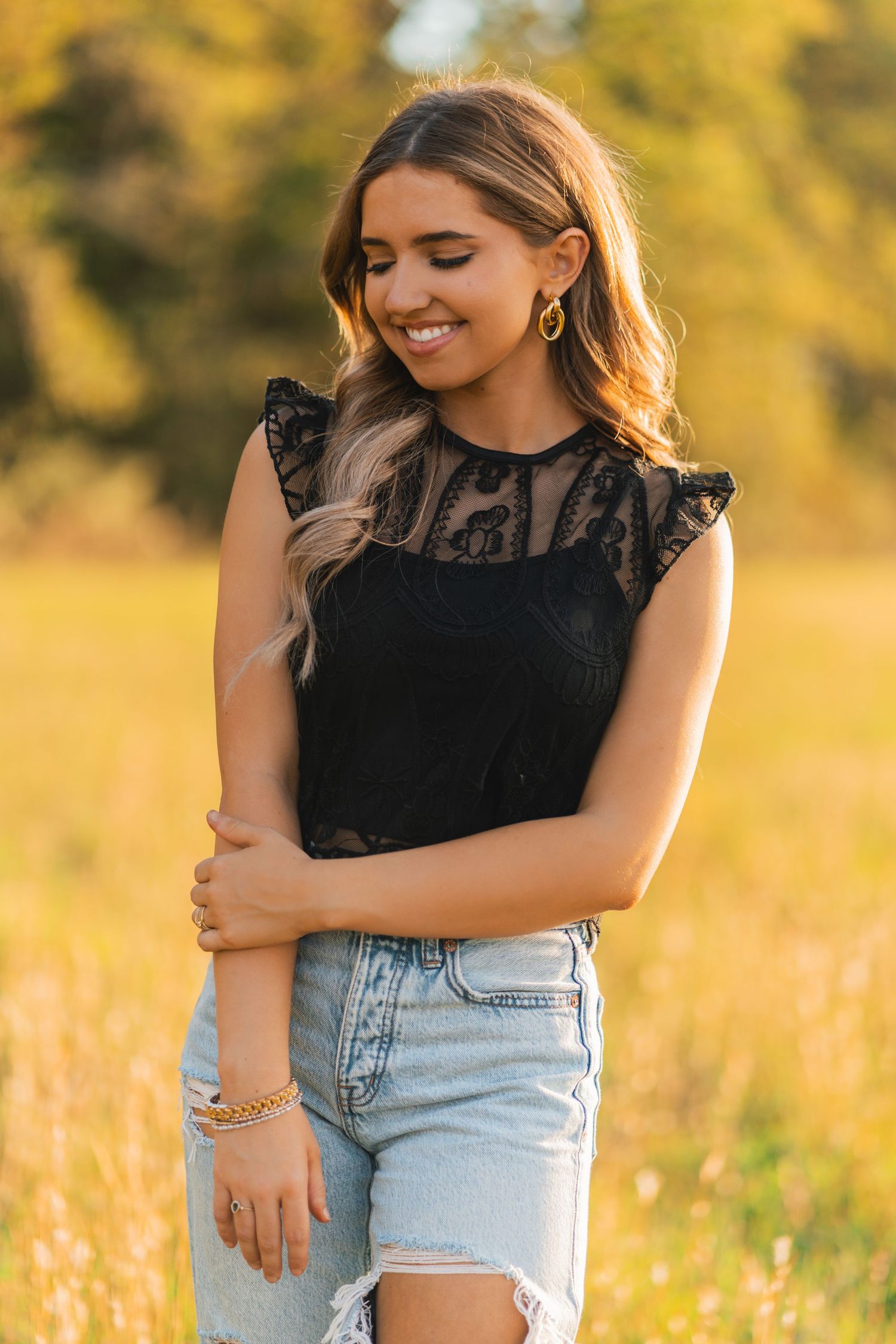 A person wearing a black lace top and distressed denim shorts poses in a sunlit golden field during sunset.