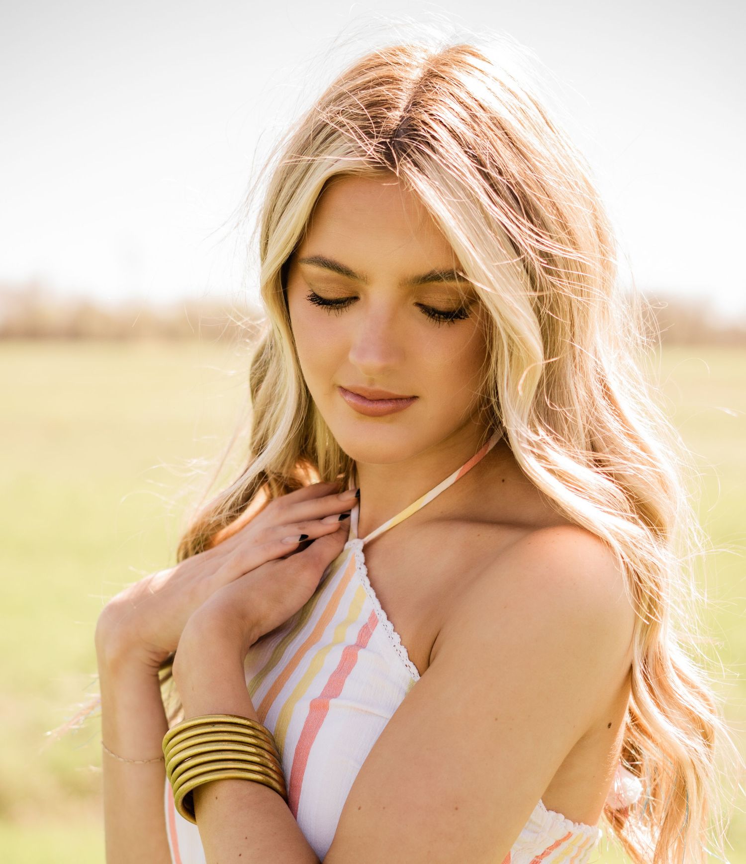 Soft sunlit portrait of a woman with wavy blonde hair wearing a white top and gold bangles in an outdoor field.