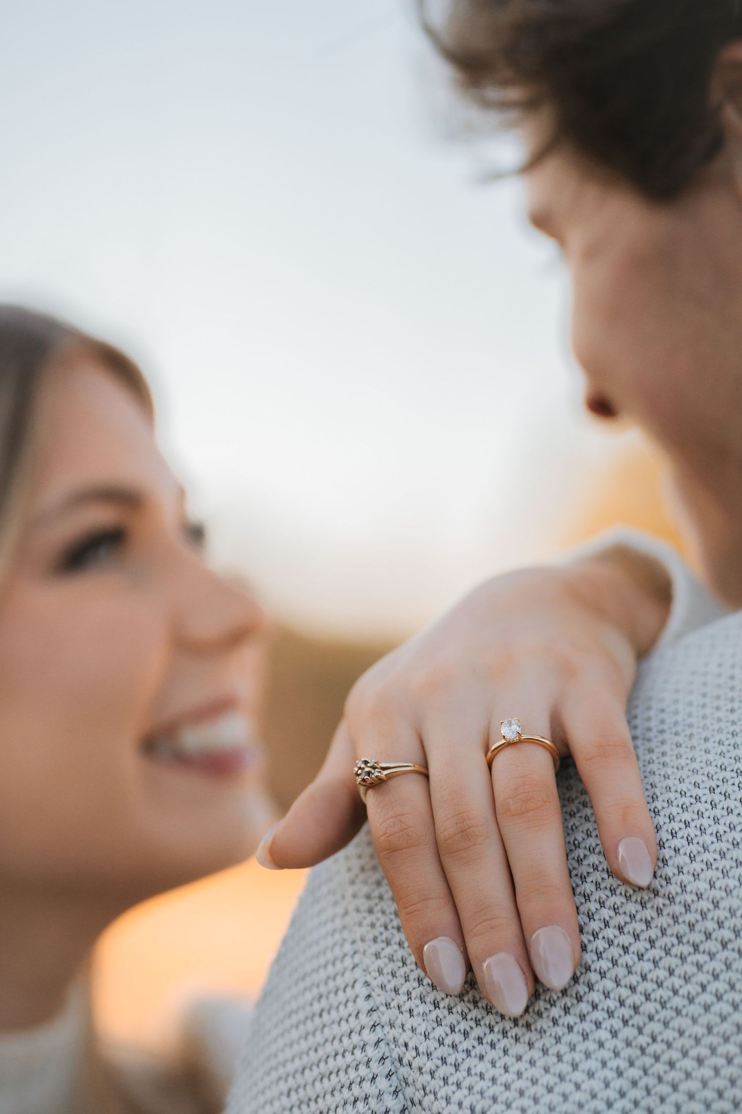 Close-up of an engagement ring on a hand resting against a gray sweater during a warm sunset moment.