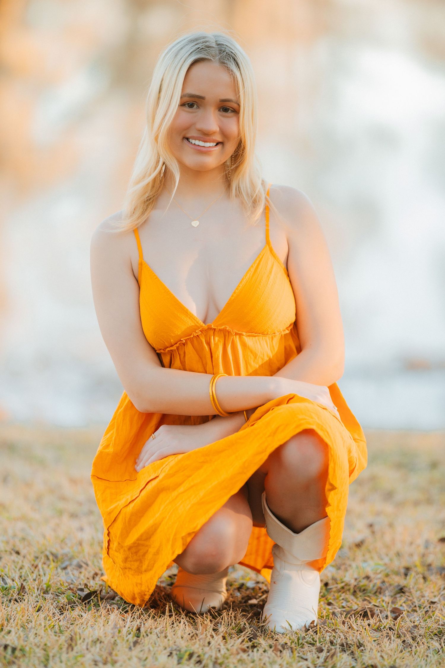 Person in flowing orange sundress and white boots poses kneeling on grass during golden hour sunset.