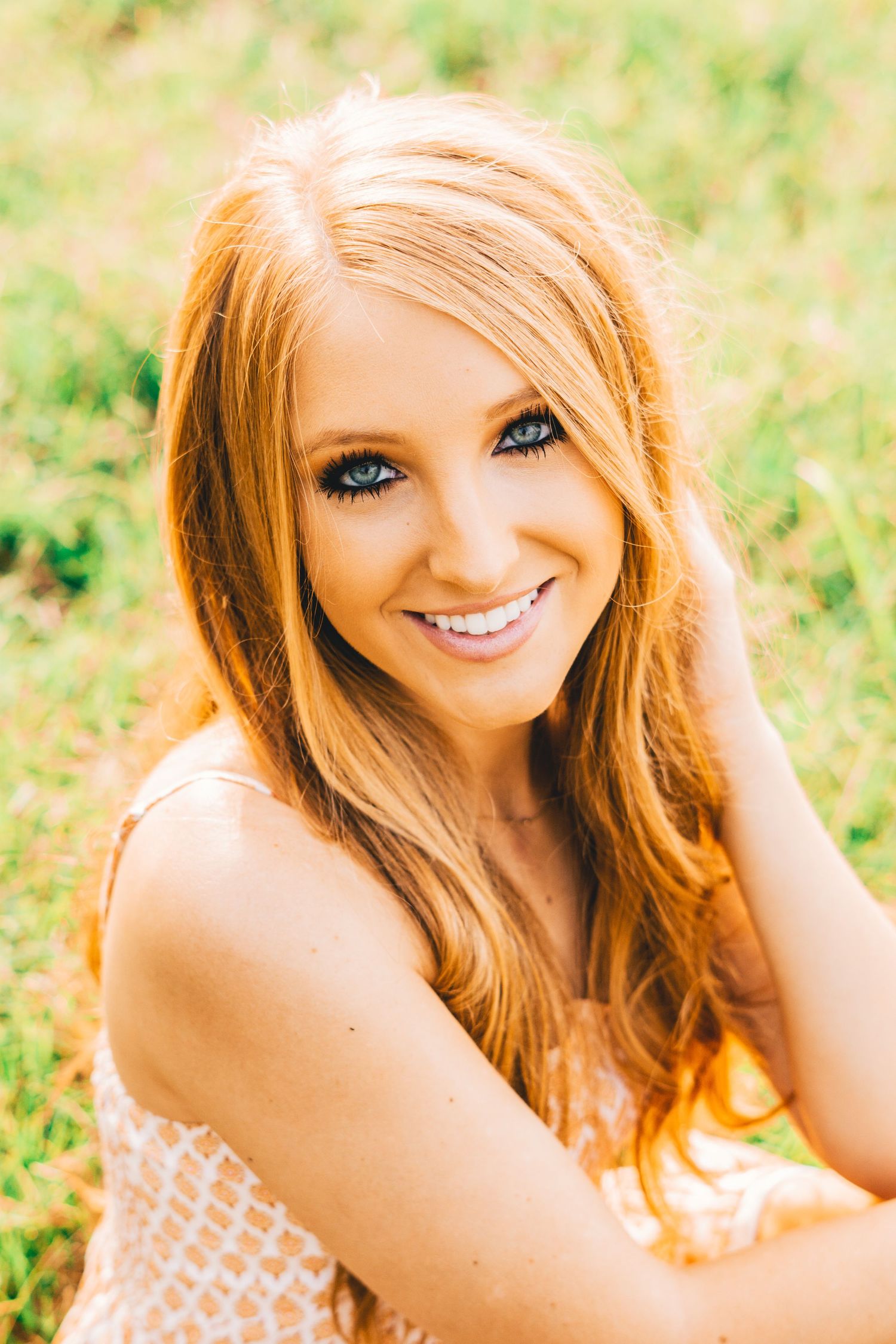 A candid outdoor portrait in natural light features bright red hair and a patterned top against a soft green background.