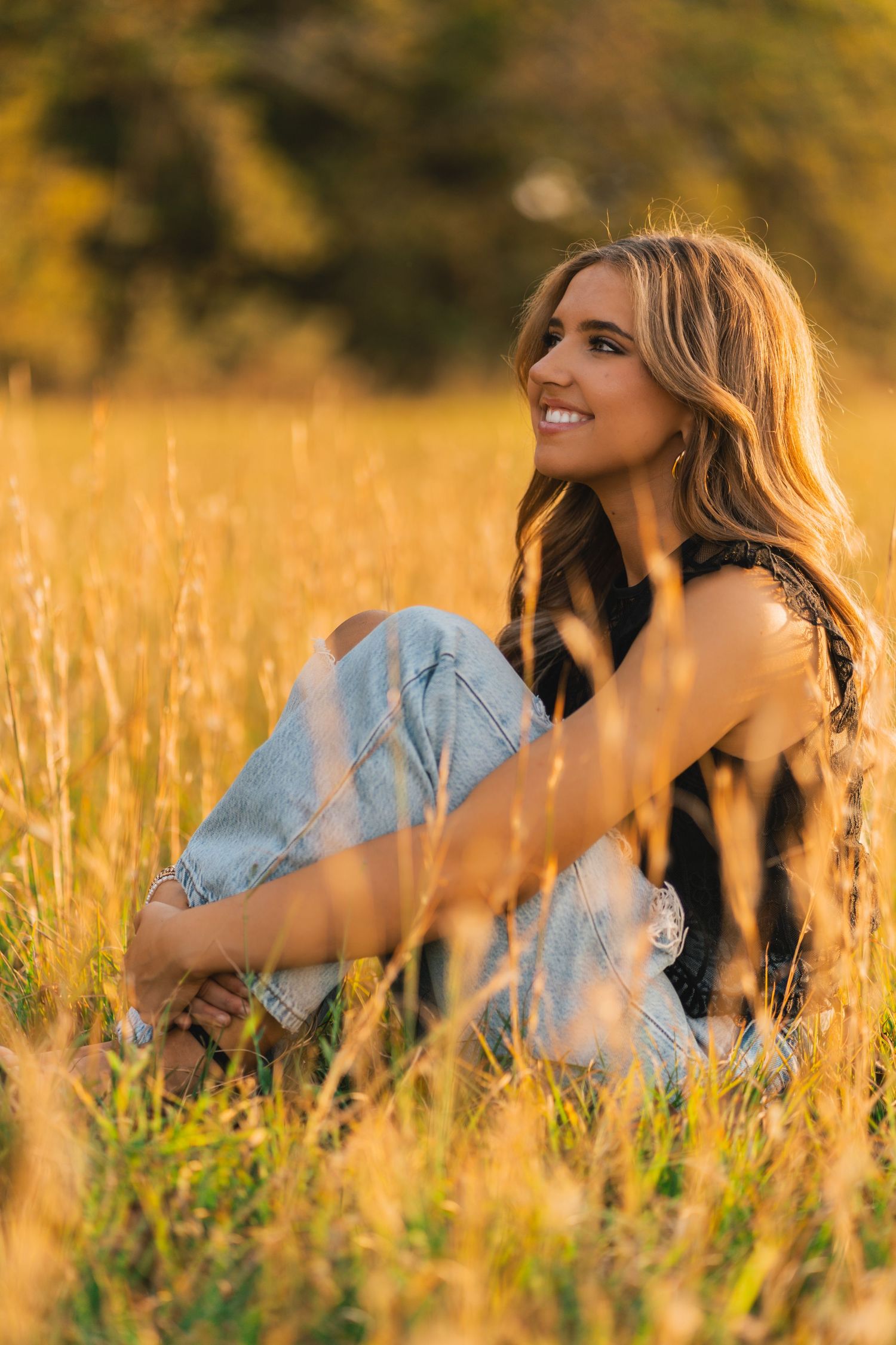 A person sits in a golden wheat field during sunset wearing jeans and a black top, smiling while looking upward.
