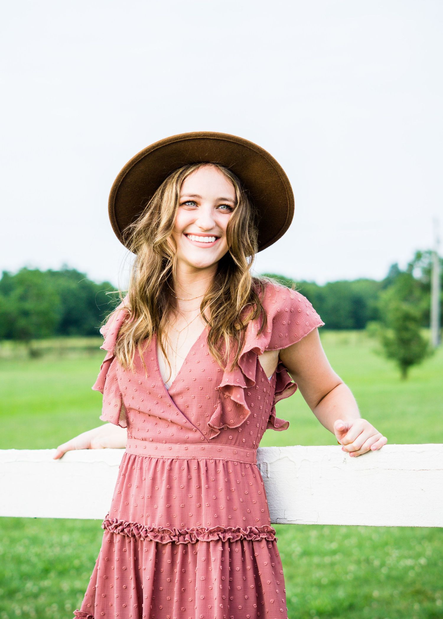 Woman in pink ruffled dress and brown hat smiles while leaning on white fence with green field in background.