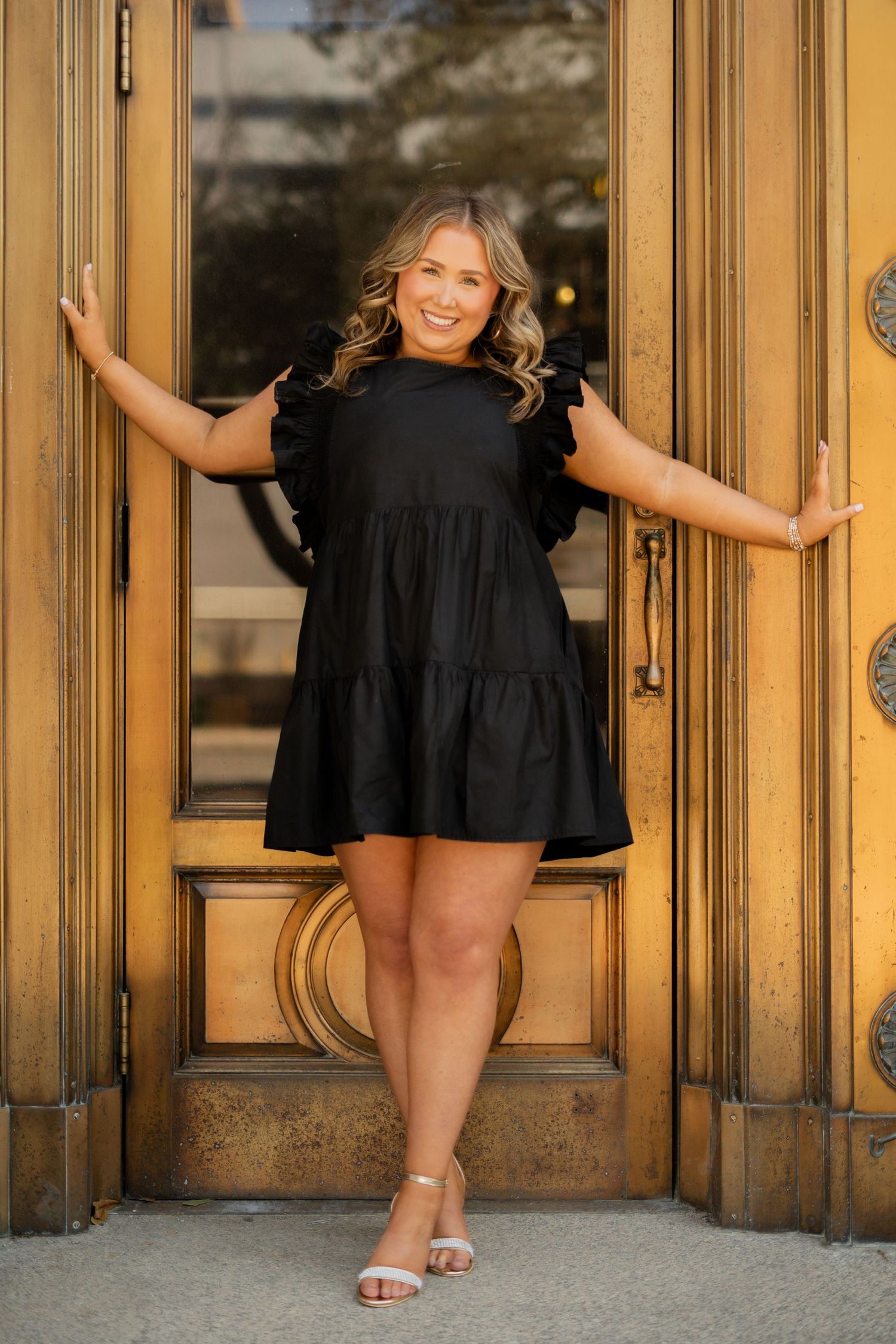 Plus size black babydoll dress with tiered ruffle design shown against ornate gold door in outdoor photoshoot setting.
