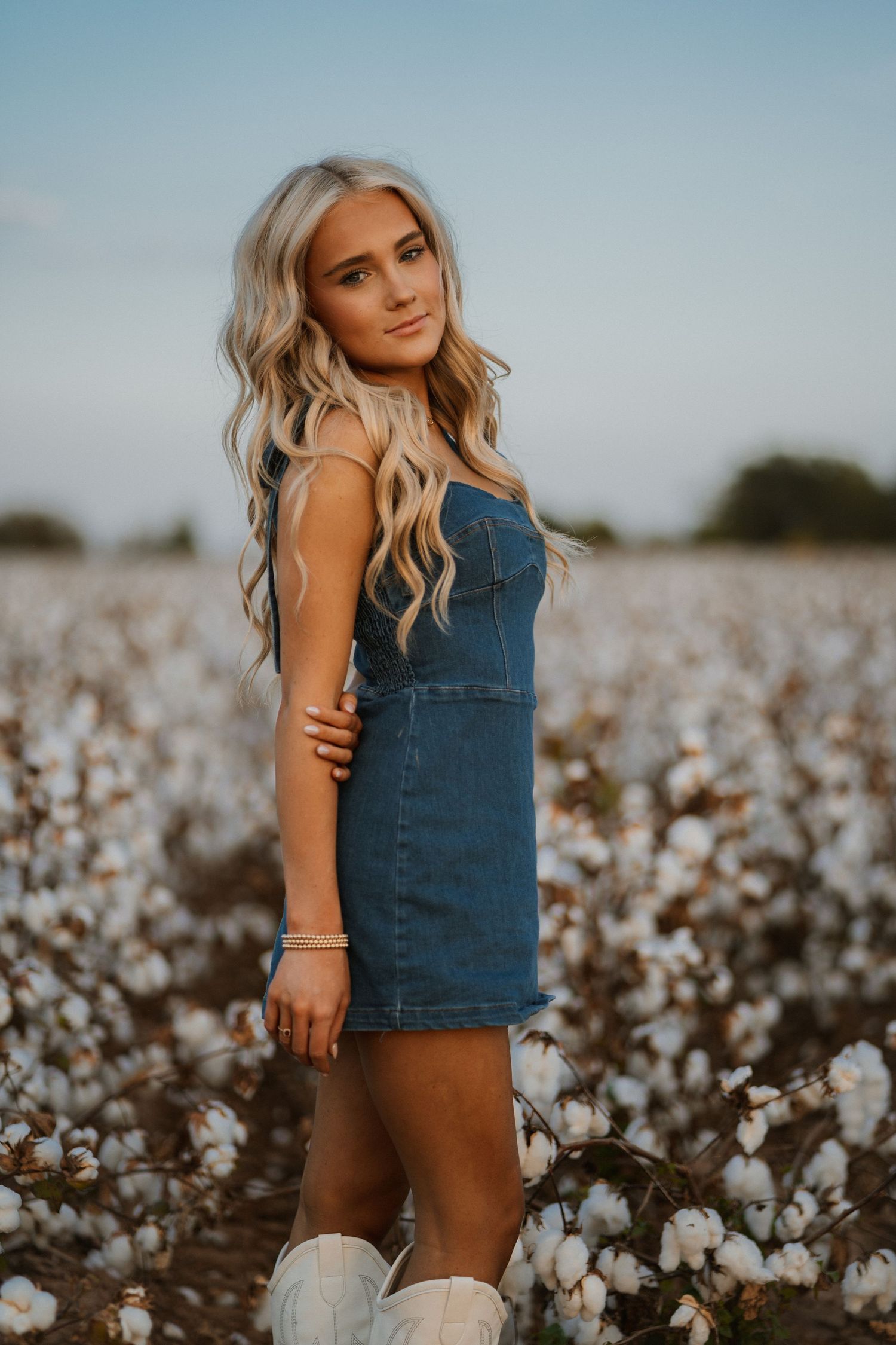 A person in a denim dress poses in a cotton field at sunset with long wavy blonde hair and white boots.