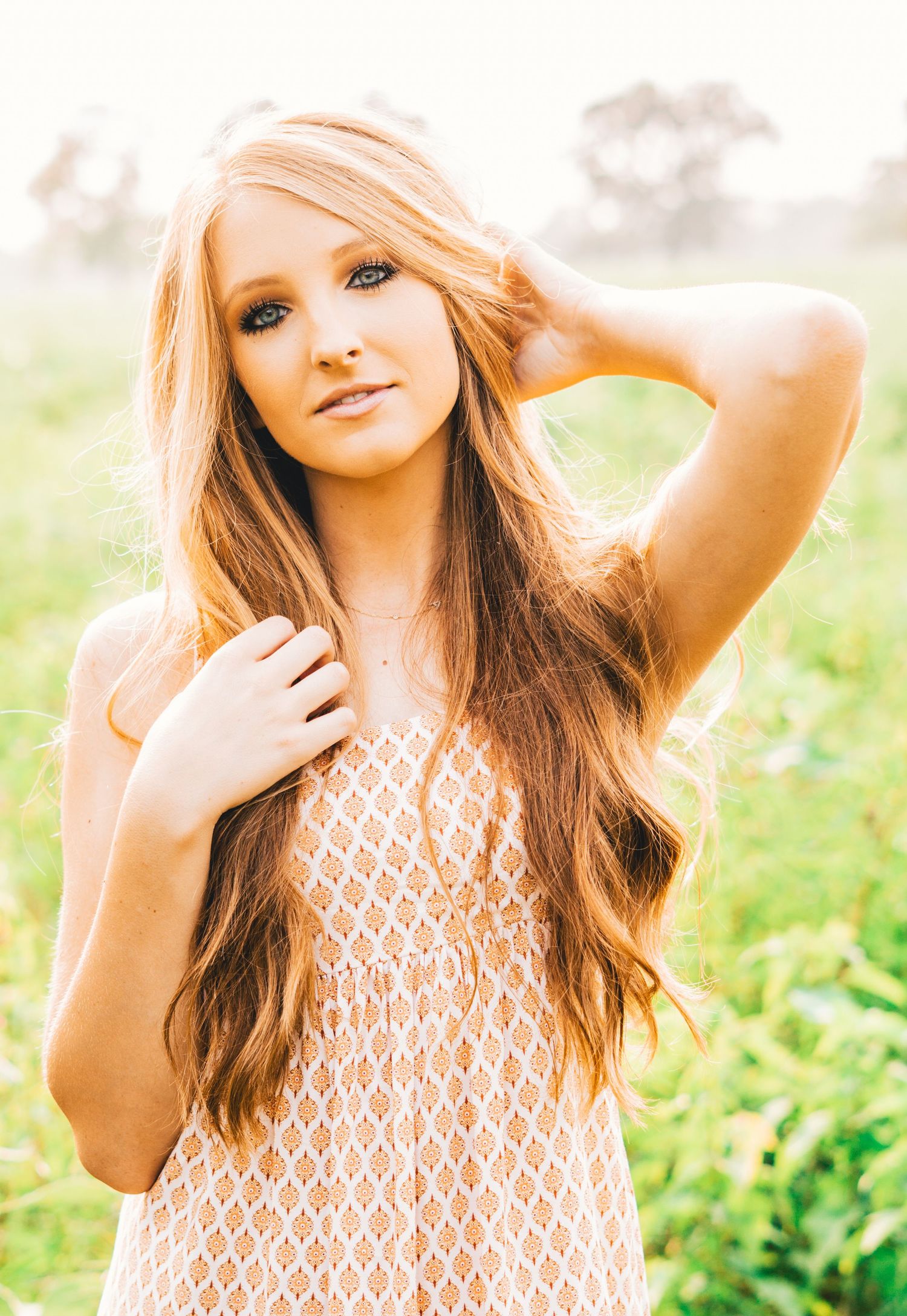 A young woman in a white lace dress poses outdoors in a natural field setting during golden hour.