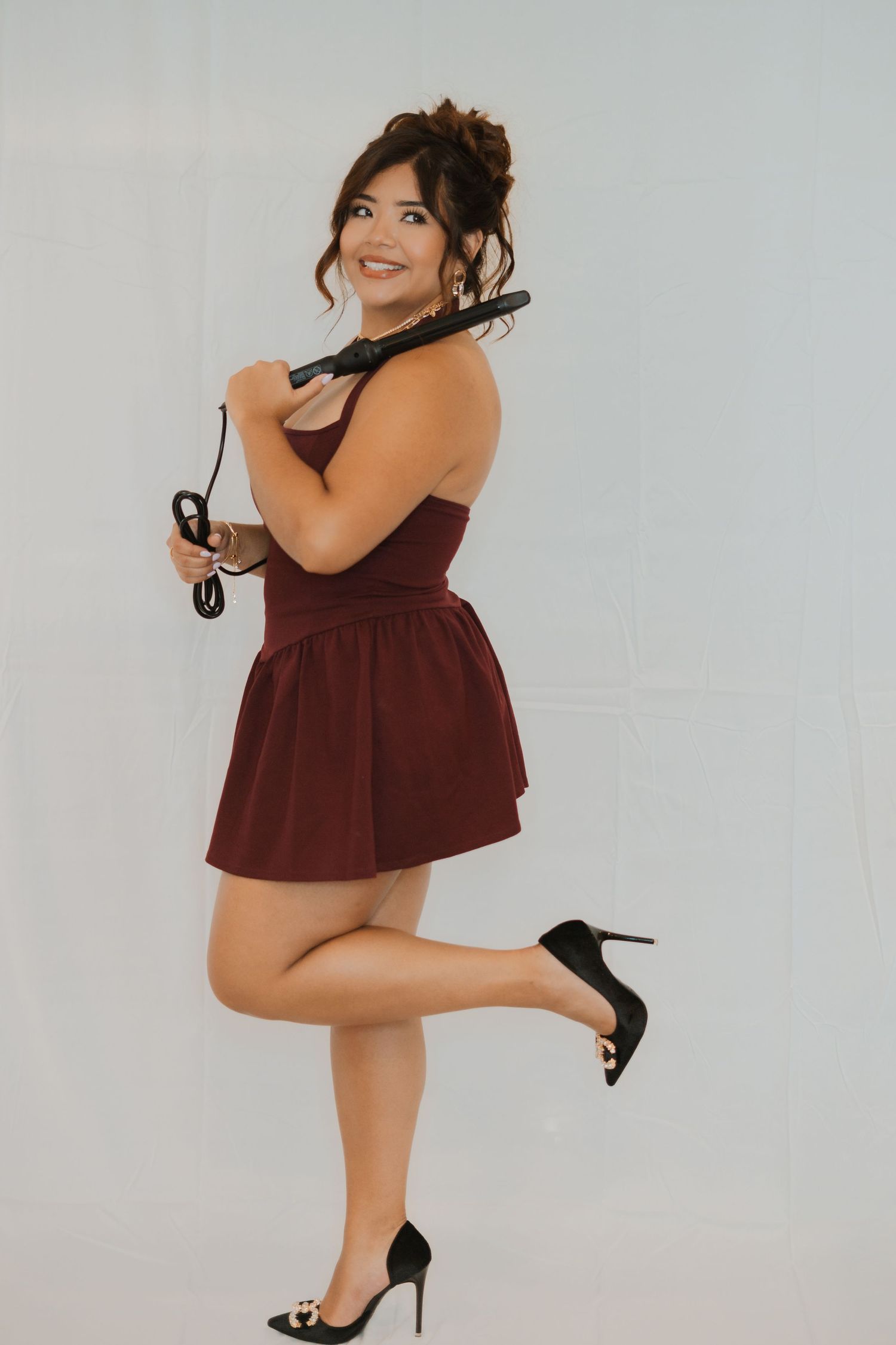 A person in a burgundy strapless dress strikes a playful pose while holding sunglasses against a light gray background.