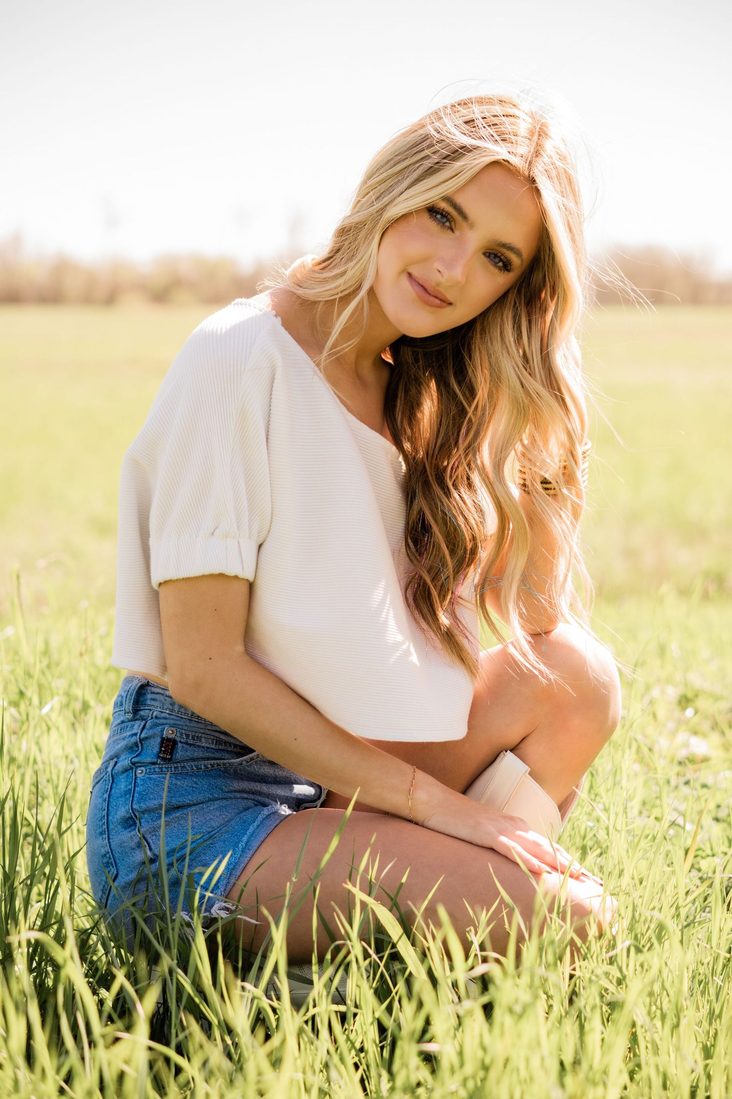 Woman in white top and denim shorts sits in sunny grass field during golden hour portrait session.