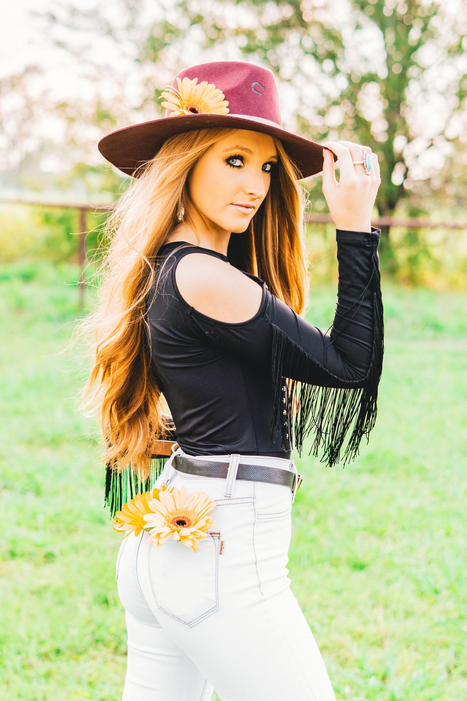 Stylish western-inspired outfit with burgundy hat, black fringe top and white jeans in sunny outdoor setting.