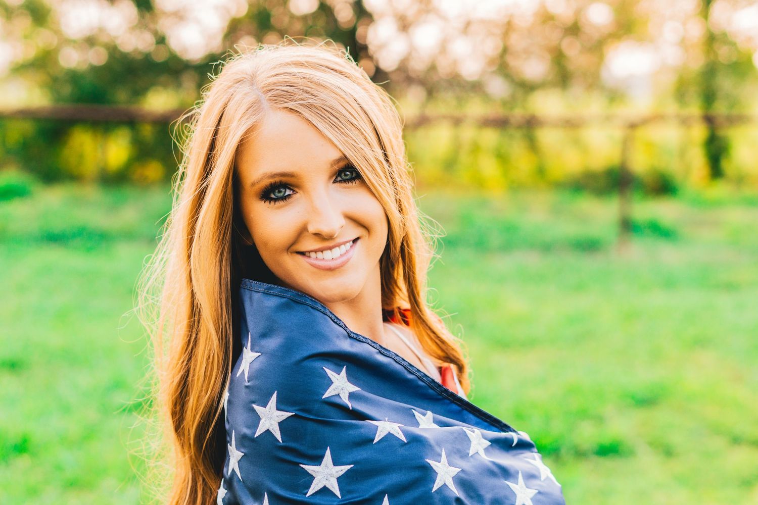Someone wrapped in American flag smiles in sunny green field with soft natural lighting and blurred background.