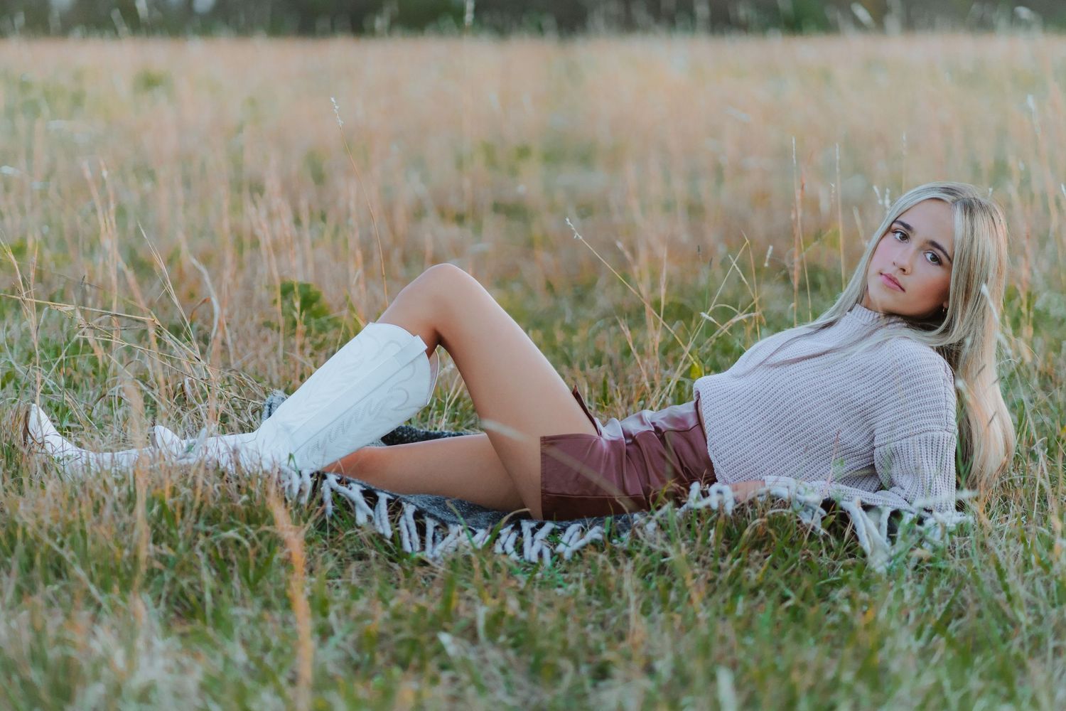 A relaxed figure in a cozy turtleneck sweater and leather shorts reclines in a golden autumn meadow wearing white boots.