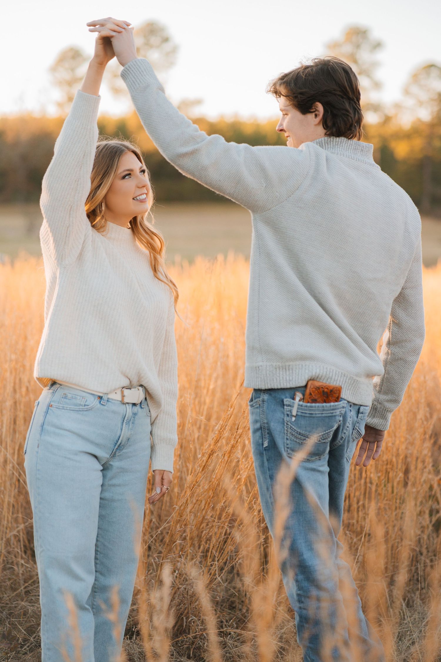 A couple dancing together in a golden field of tall grass at sunset wearing matching light colored sweaters and jeans.