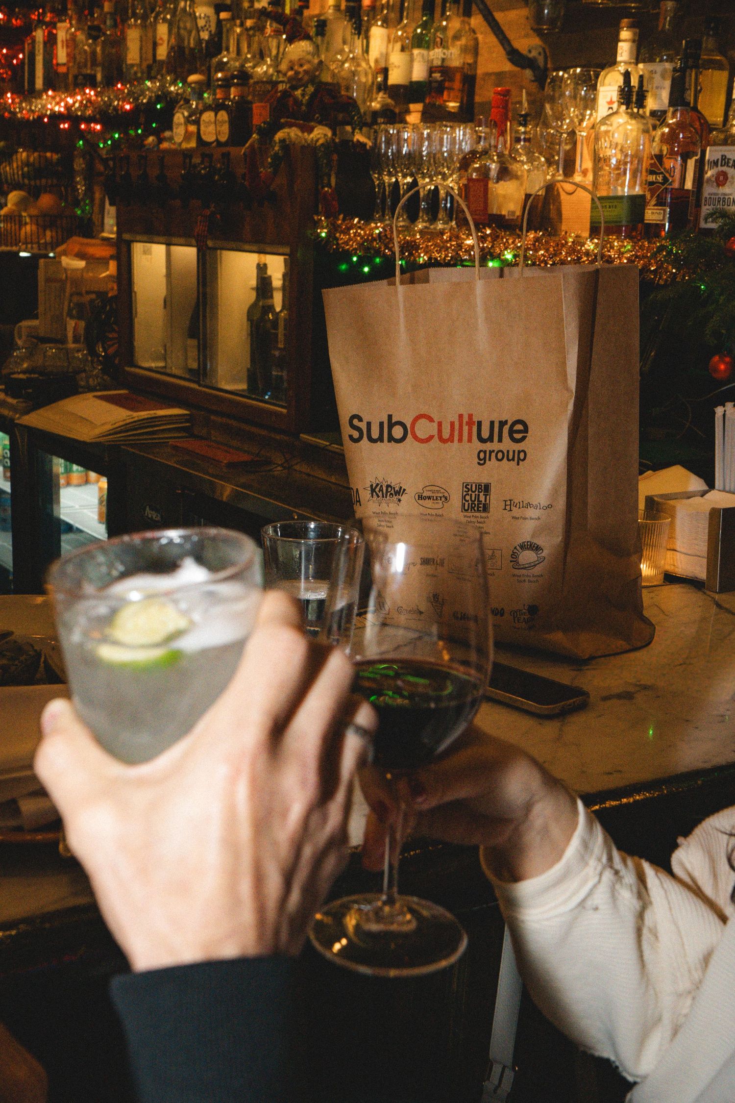 Two hands clinking cocktail glasses at a bar with a SubCulture Group paper bag and bottles in background.