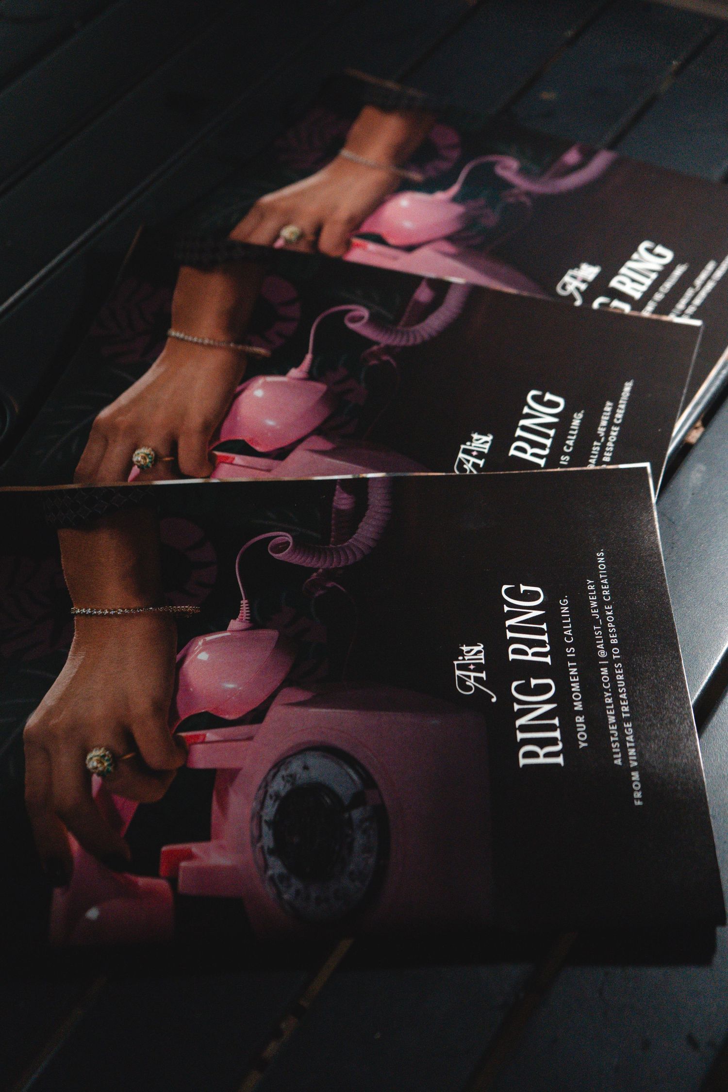 Stack of Ring Ring event programs featuring a pink rotary phone and a hand with rings on a dark wooden surface.