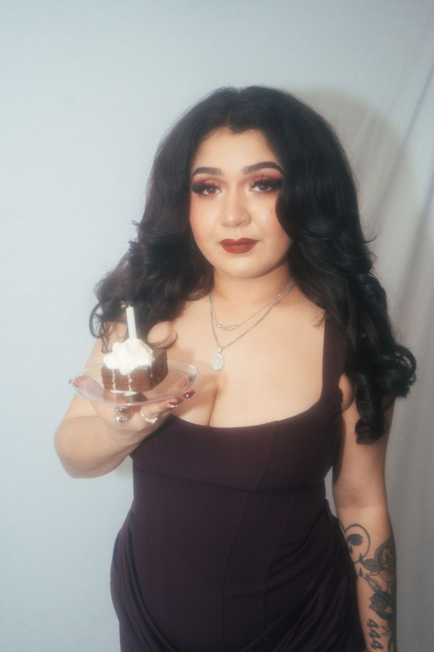 A tattooed woman in a dark dress holds a cupcake with a candle while posing.