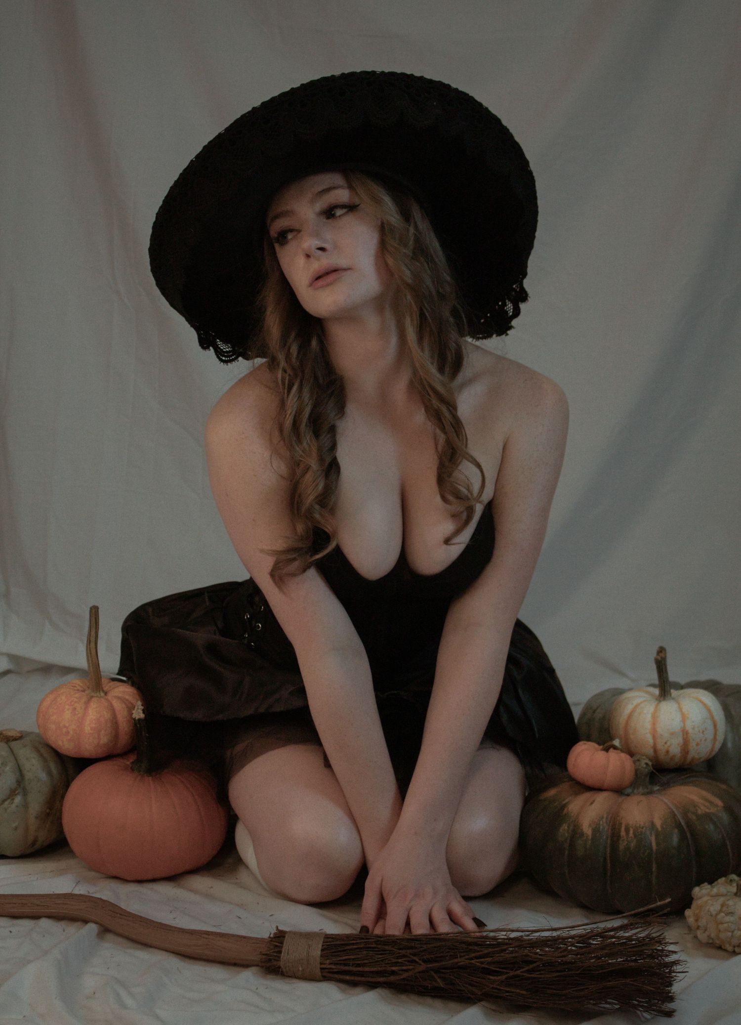 Woman in witch costume with black hat sitting among colorful pumpkins and a broom in a Halloween photoshoot.