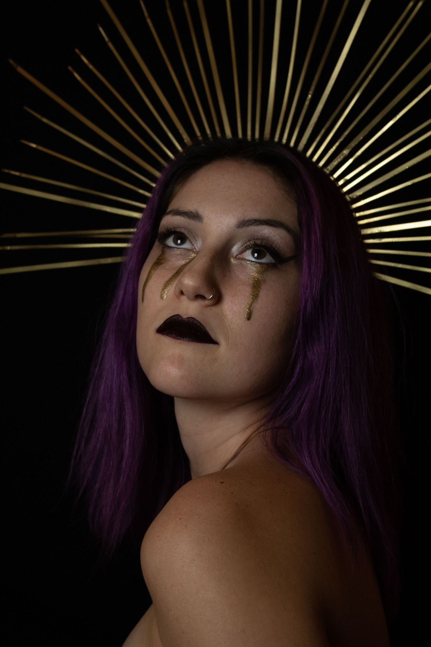 Portrait of woman with purple hair wearing gold halo crown with metallic tear streaks on dark background.