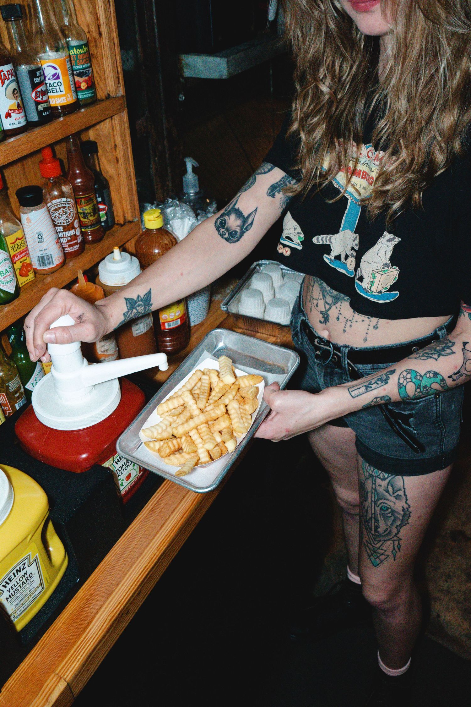 Tattooed woman dispensing sauce over waffle fries on a metal tray at a bar condiment station.