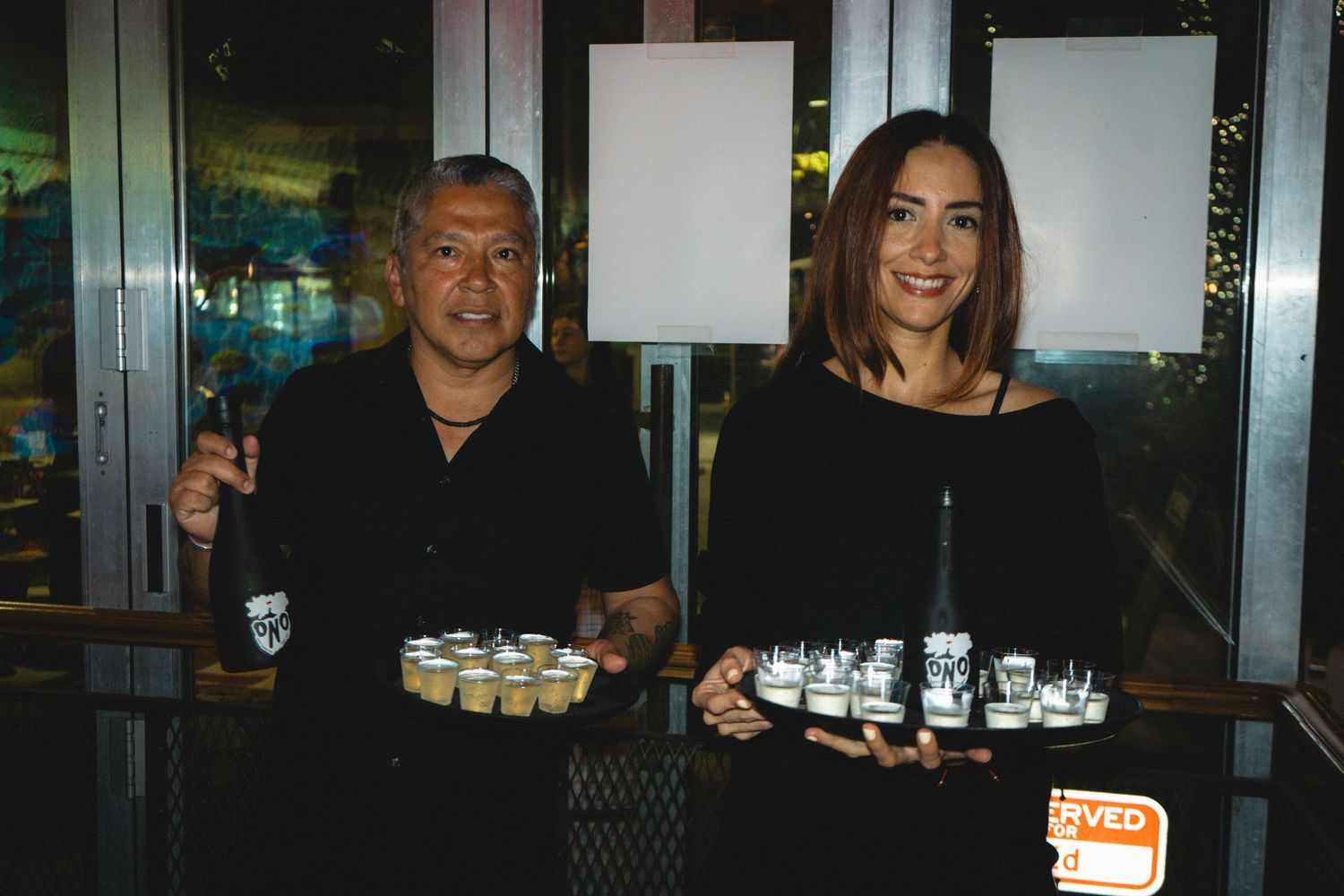Two people smiling while holding trays of sake shots and bottles at a Sake ONO event.
