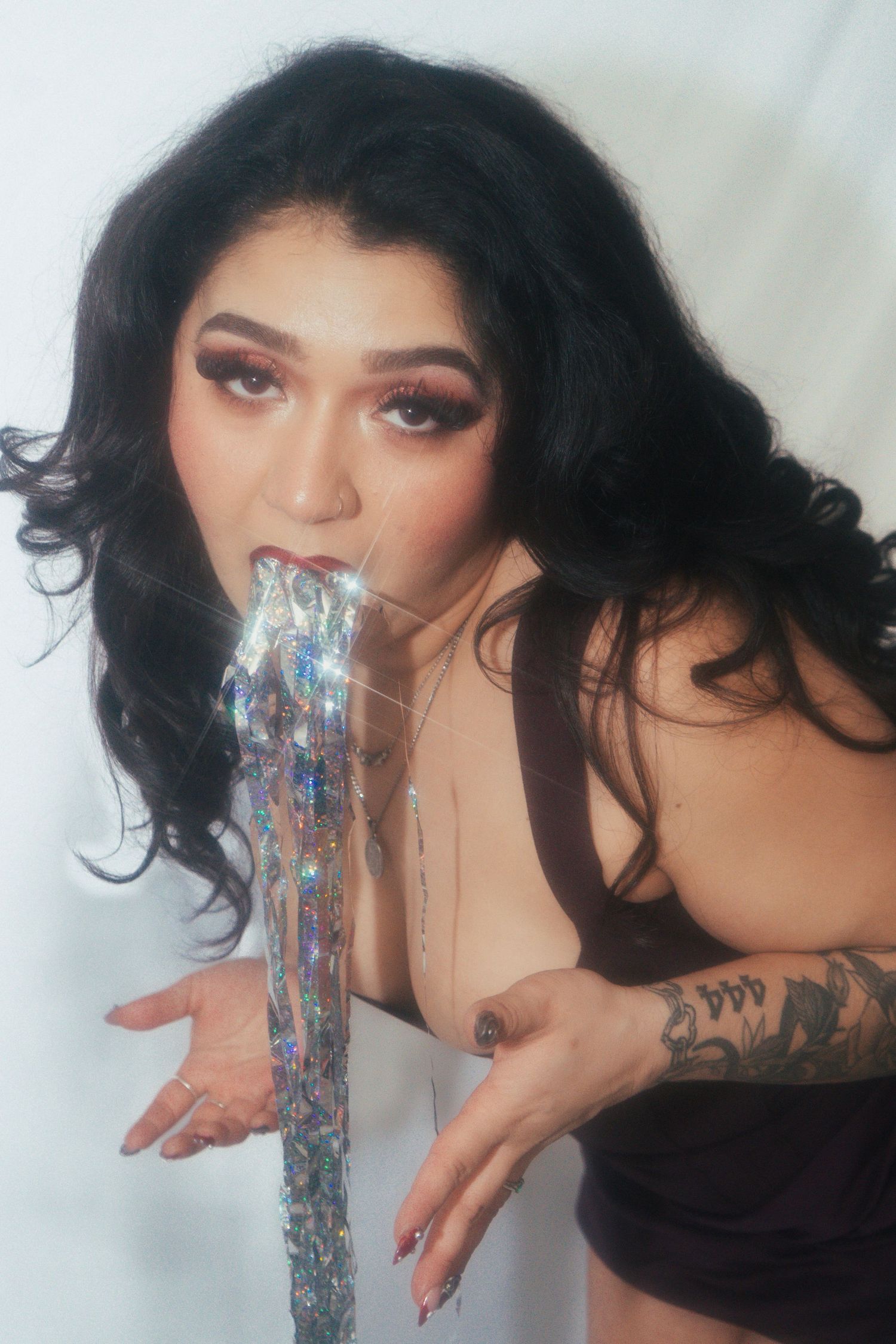 Woman with dark wavy hair and bold makeup posing with holographic tinsel against white background.