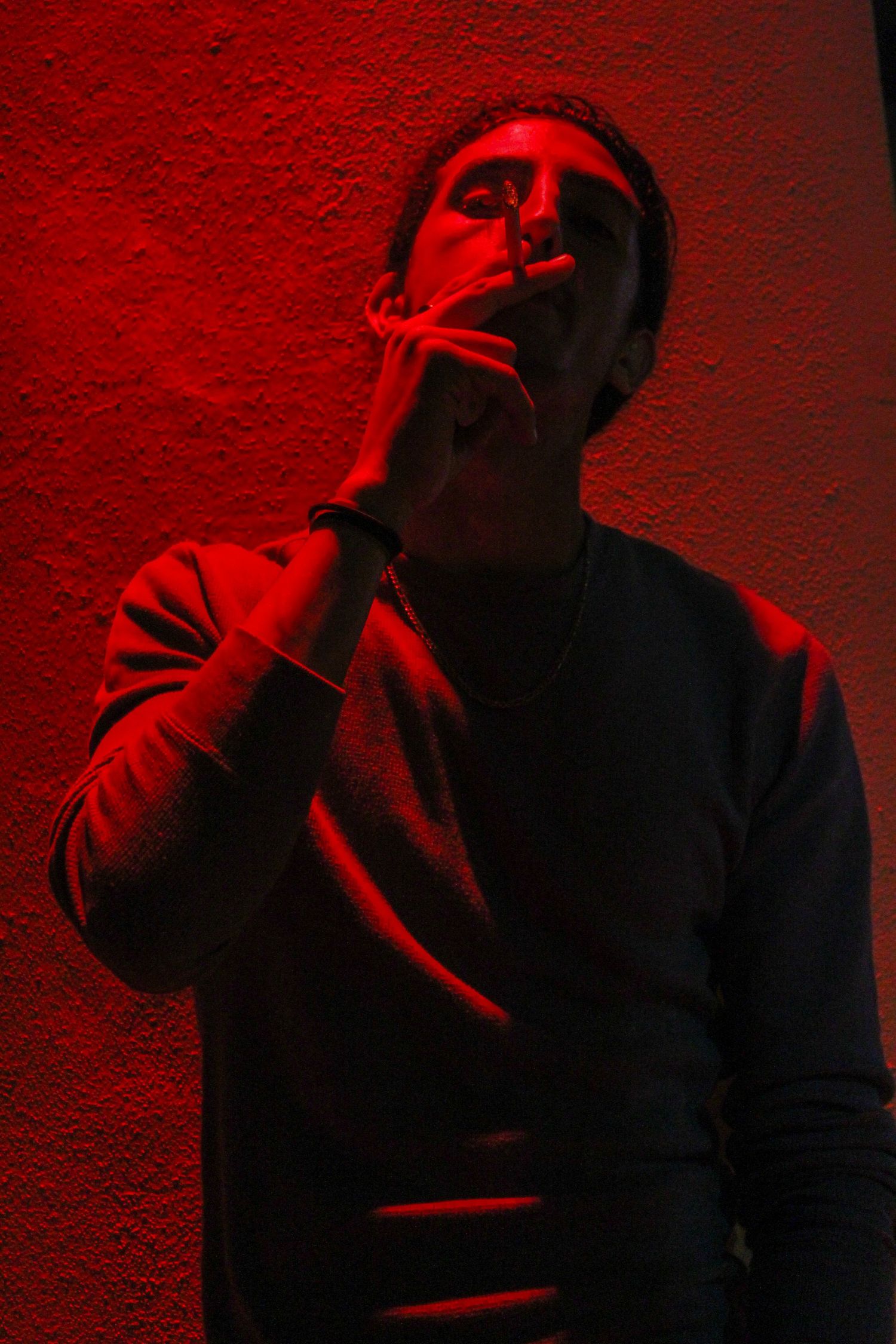Man in grey sweatshirt smoking a cigarette while leaning against textured wall under atmospheric red lighting.