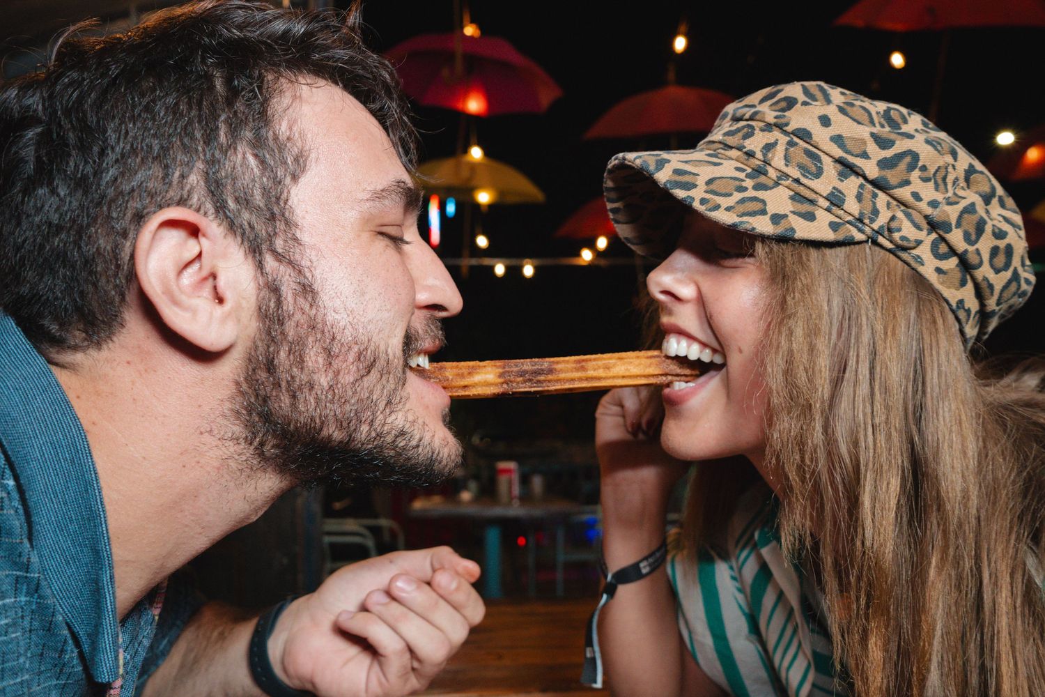 A couple playfully sharing a churro at a festively lit Mexican restaurant with colorful hanging lights.