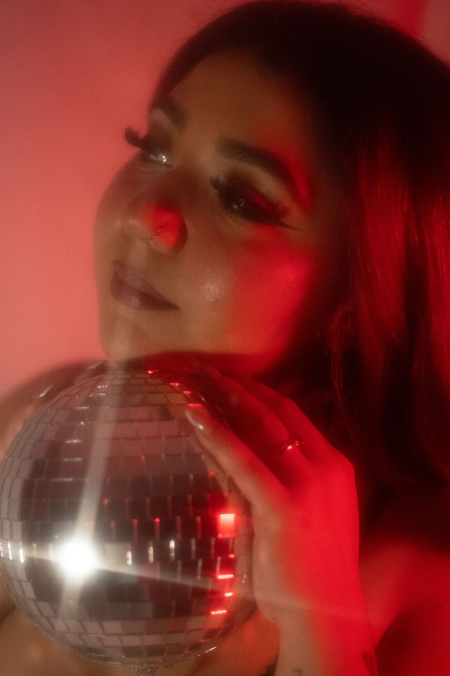 Woman with bold red eye makeup resting cheek on silver disco ball in red light.