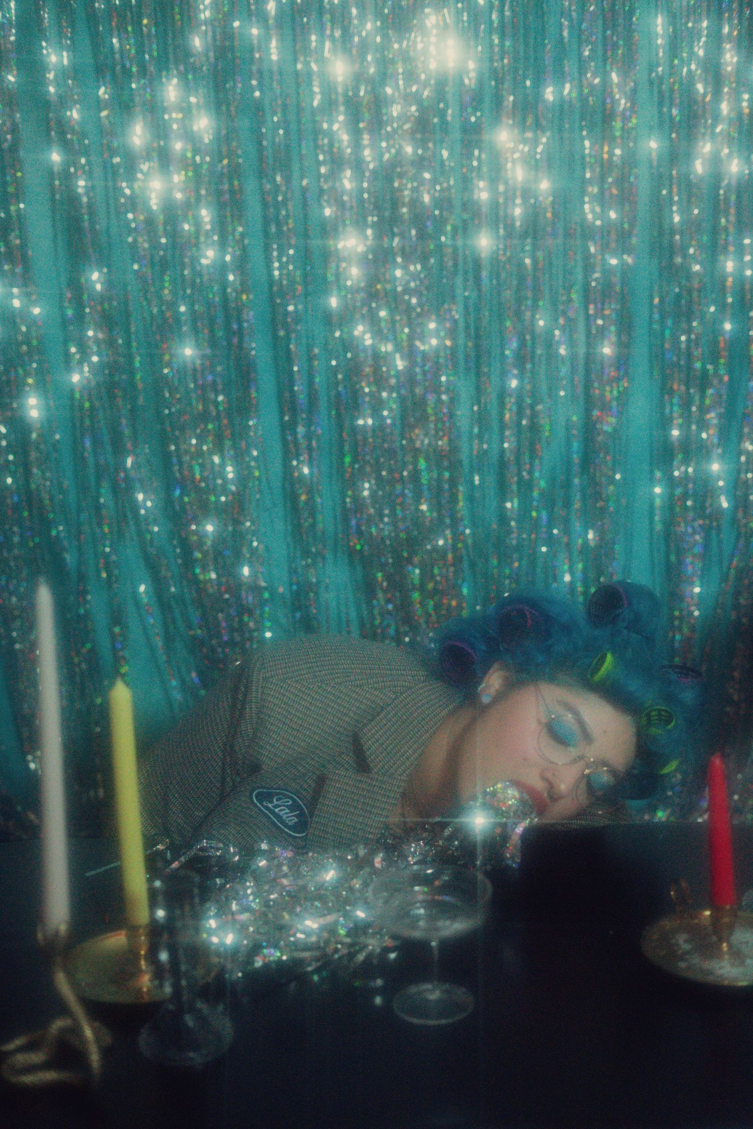 Woman with blue hair lies among candles and glitter on a table with a sparkly teal backdrop behind her.