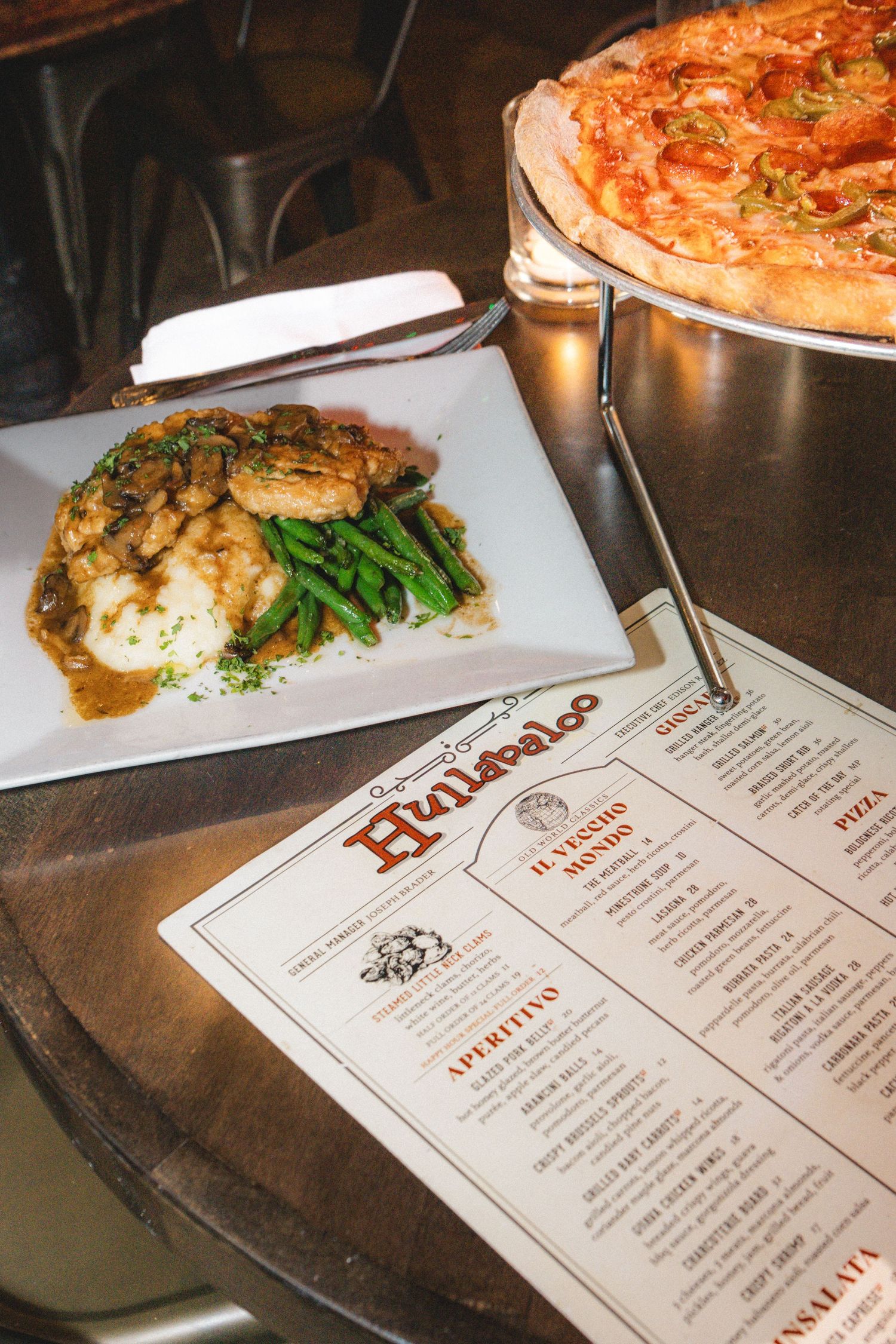 Italian restaurant table with chicken entree, green beans, pizza on stand, and Hunipalo menu card.