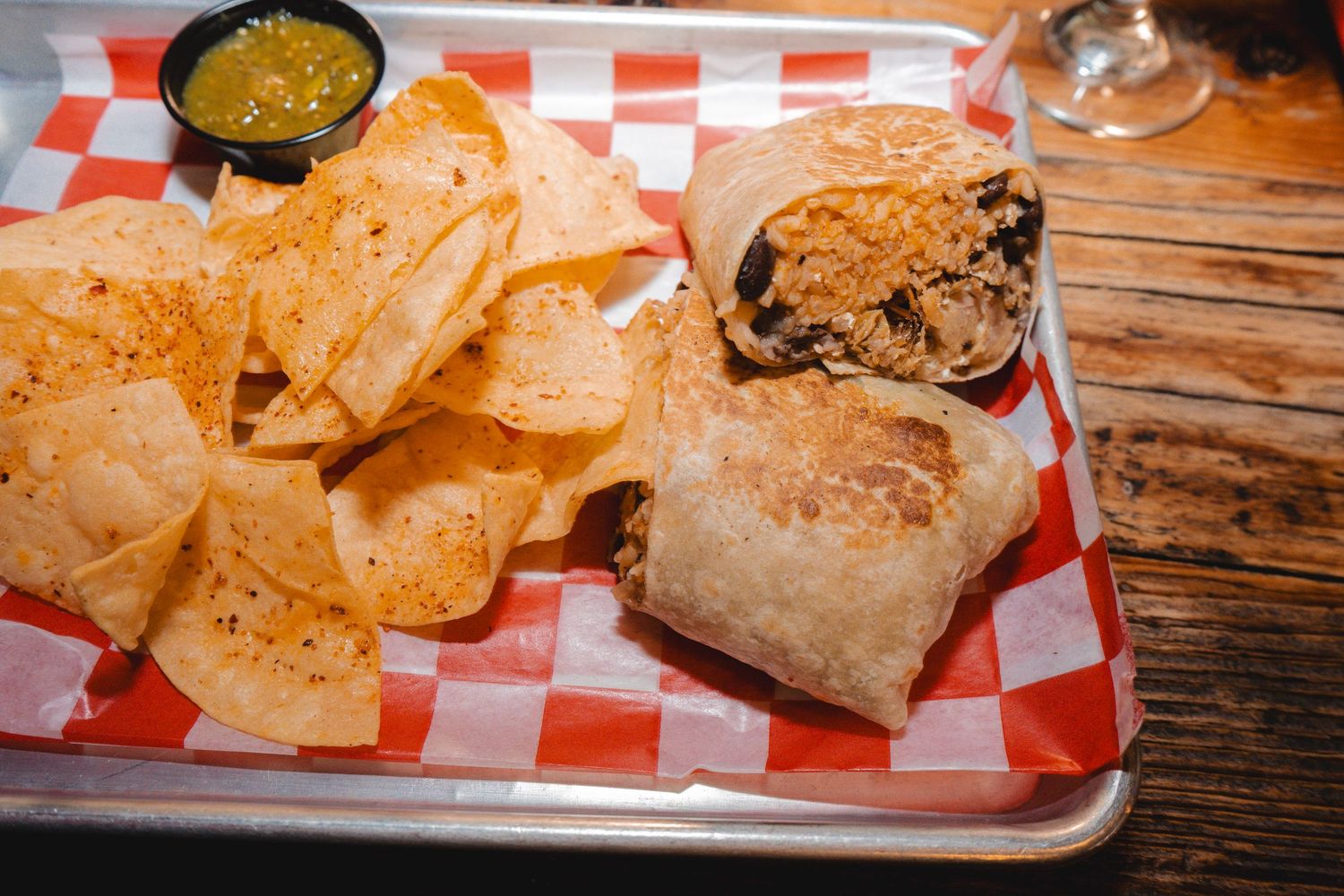 A hearty burrito cut in half served with seasoned tortilla chips and green salsa on a metal tray.