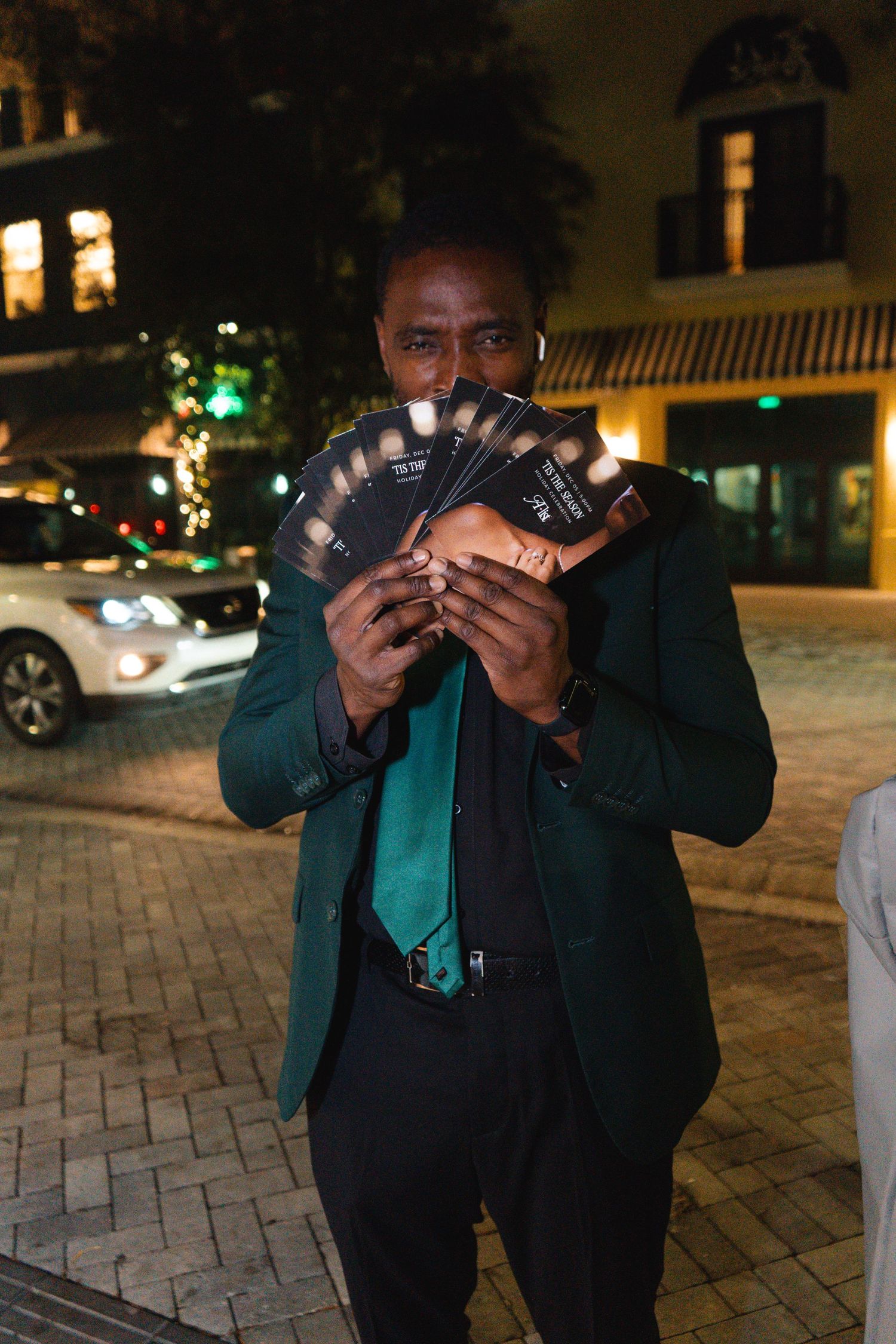 Stylish man in green blazer holding fanned-out event tickets outside at night on a city street.