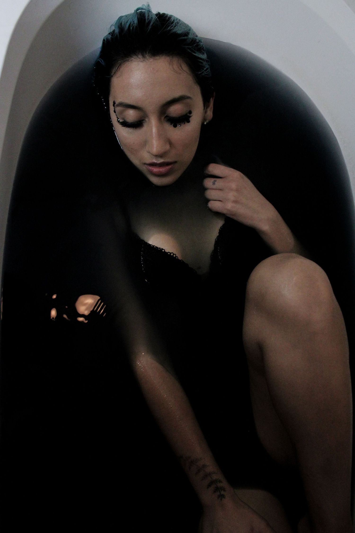 A woman reclines in a bathtub filled with dark water, wearing black lingerie with a tattoo visible on her arm.