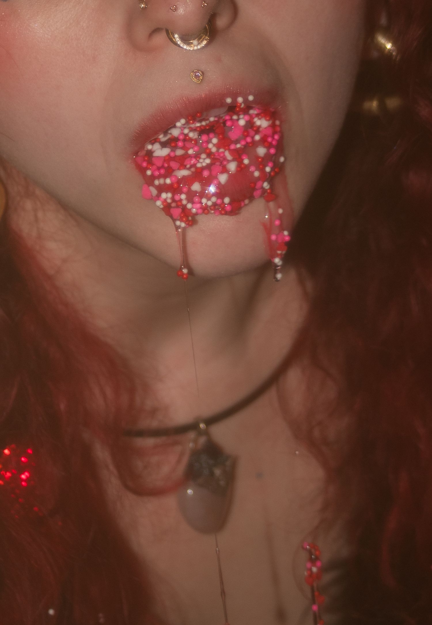 Close-up of lips with pink and white sprinkles dripping down chin, with nose ring and crystal necklace visible.