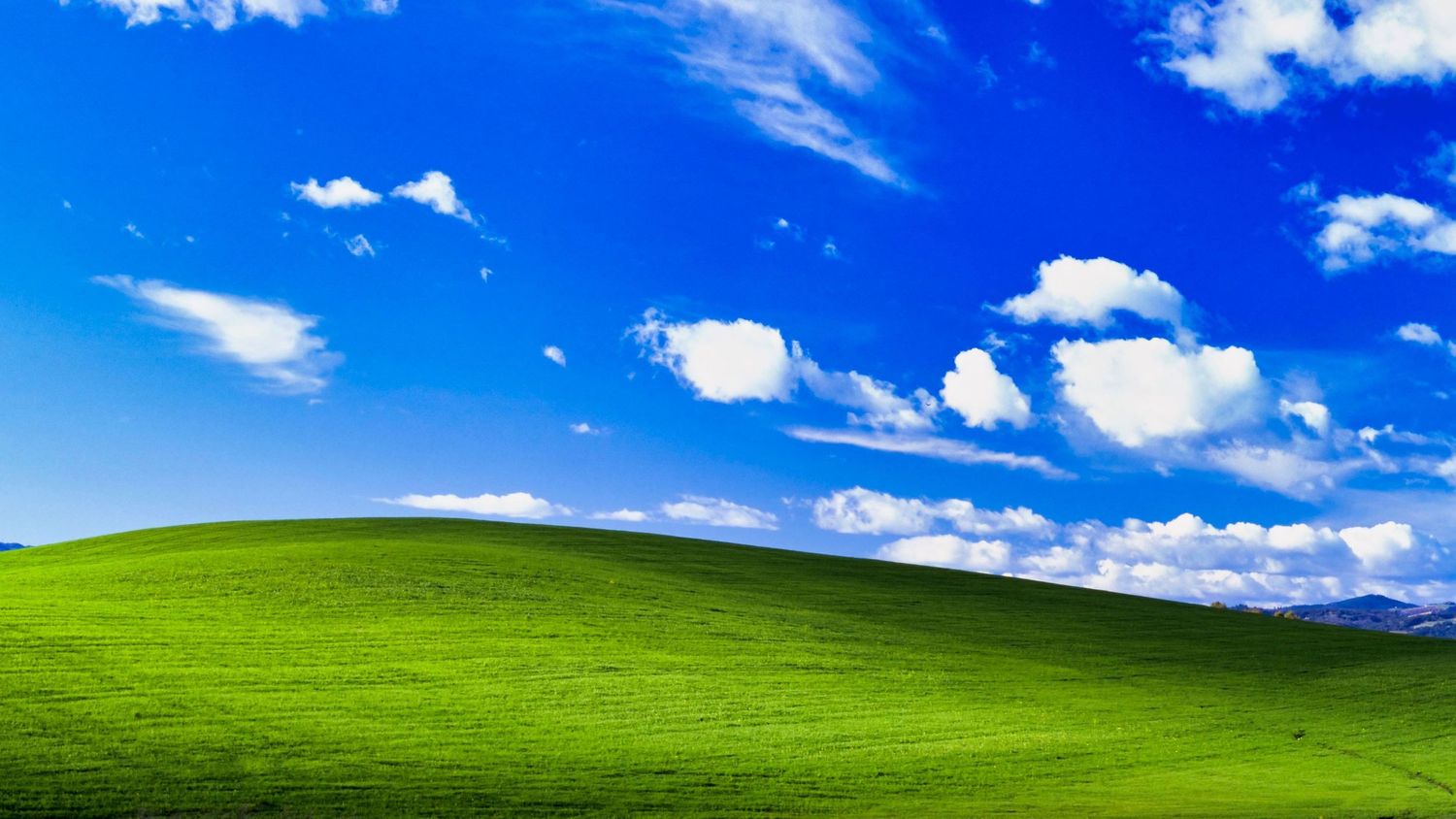 Lush green rolling hills under a bright blue sky with fluffy white clouds, resembling the classic Windows XP Bliss wallpaper.