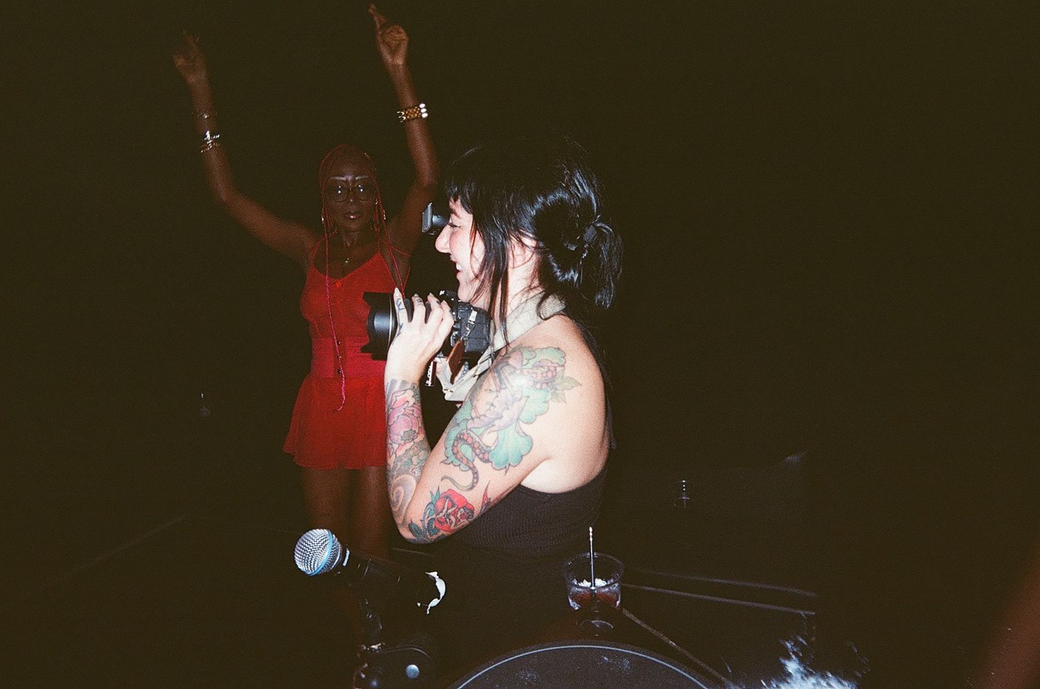 Tattooed female singer performs on stage while a woman in red dress dances behind her at a concert.
