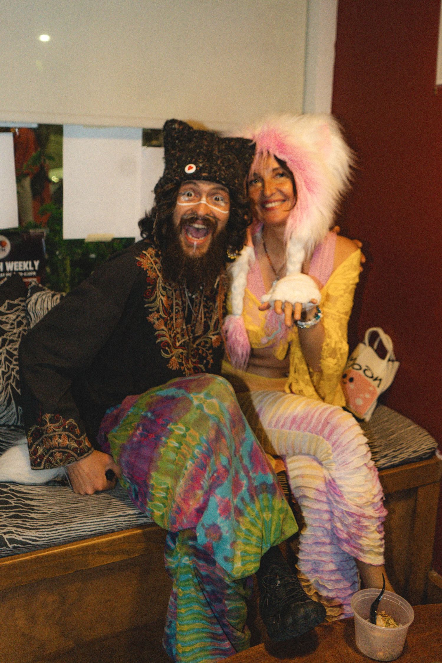 Two people in colorful tie-dye and fur costumes pose together at a Halloween party, smiling and holding a white object.