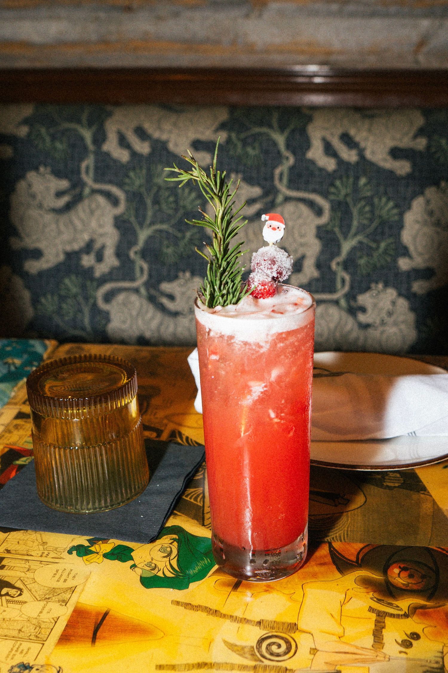 A vibrant red holiday cocktail with rosemary sprig and Santa garnish in a highball glass at a restaurant booth.