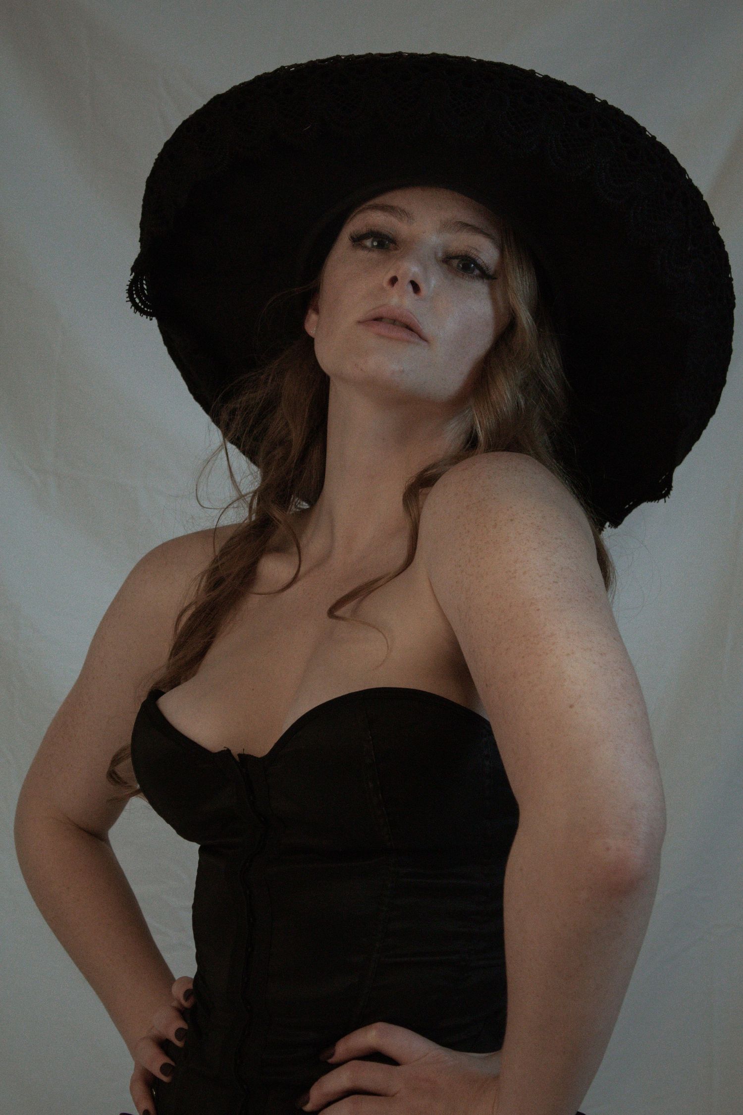 Woman in black strapless corset and wide-brim black hat posing against a light fabric backdrop.