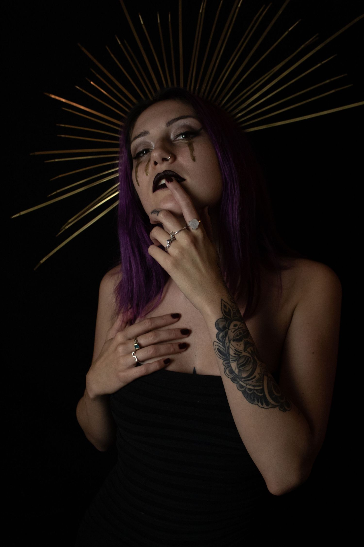 Woman with purple hair and gold spike halo crown posing with tattooed hands near face on dark background.