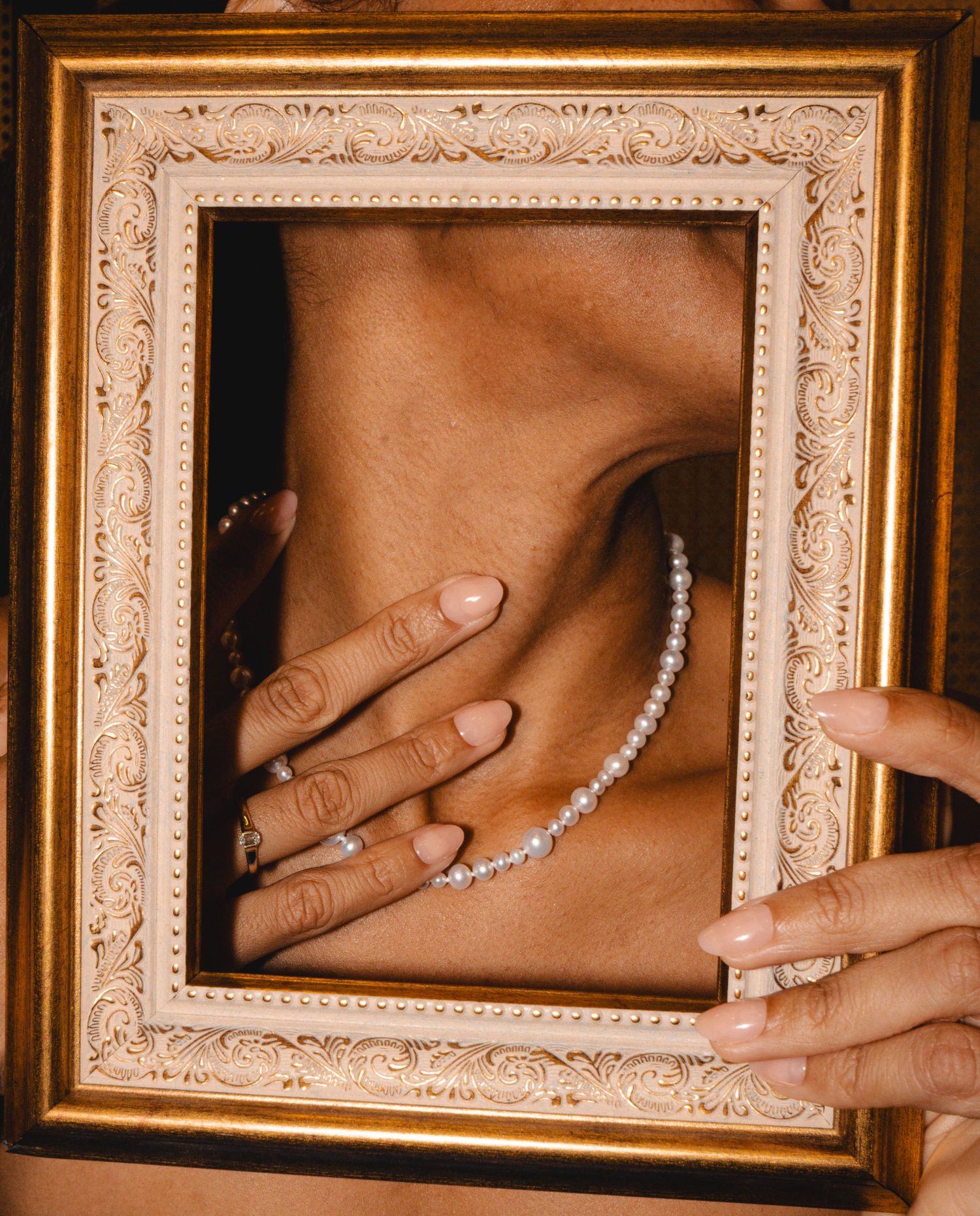 A pearl necklace and manicured hand displayed within an ornate gold picture frame.