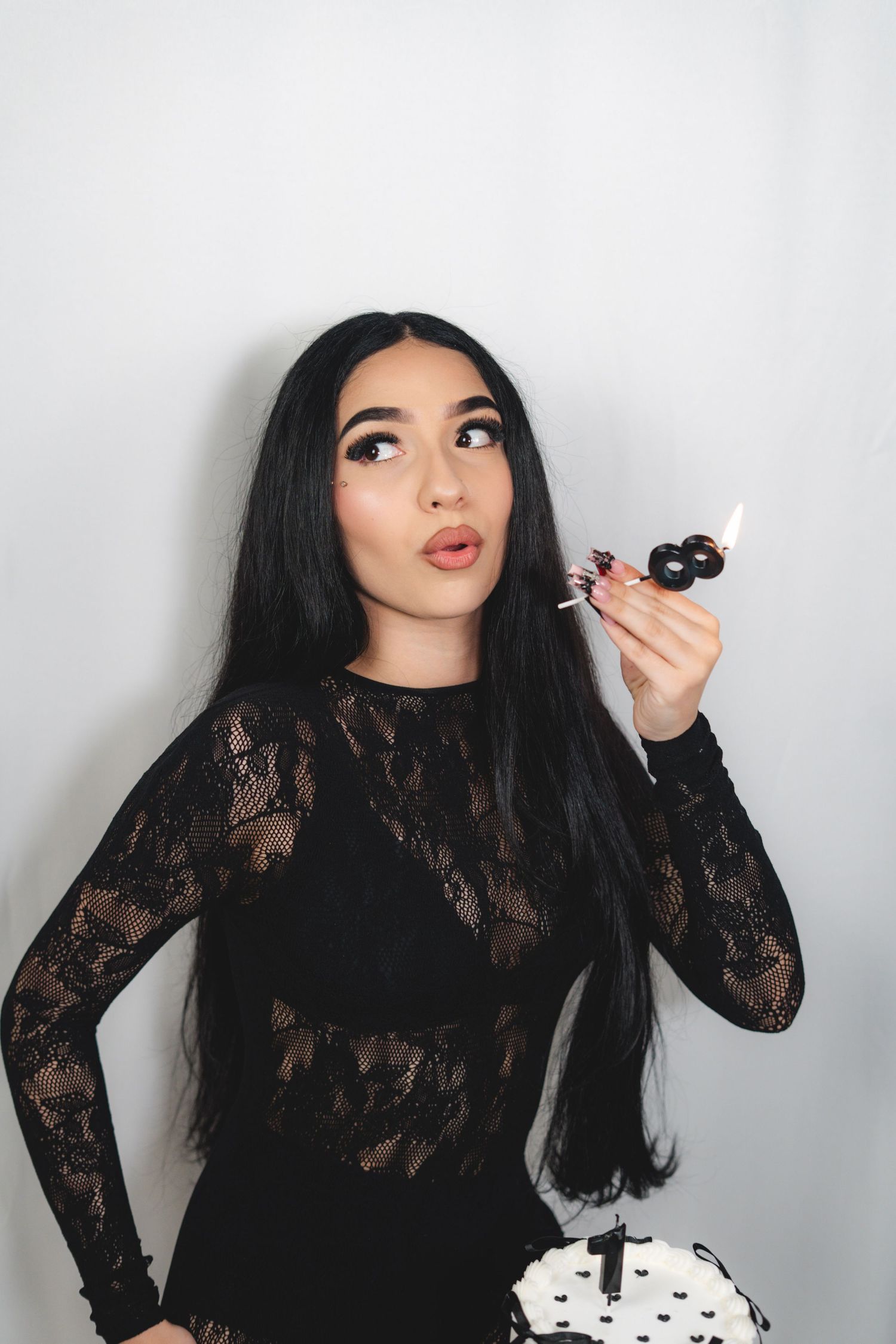 Young woman in black lace dress holding a lighter near candles on an 18th birthday cake.