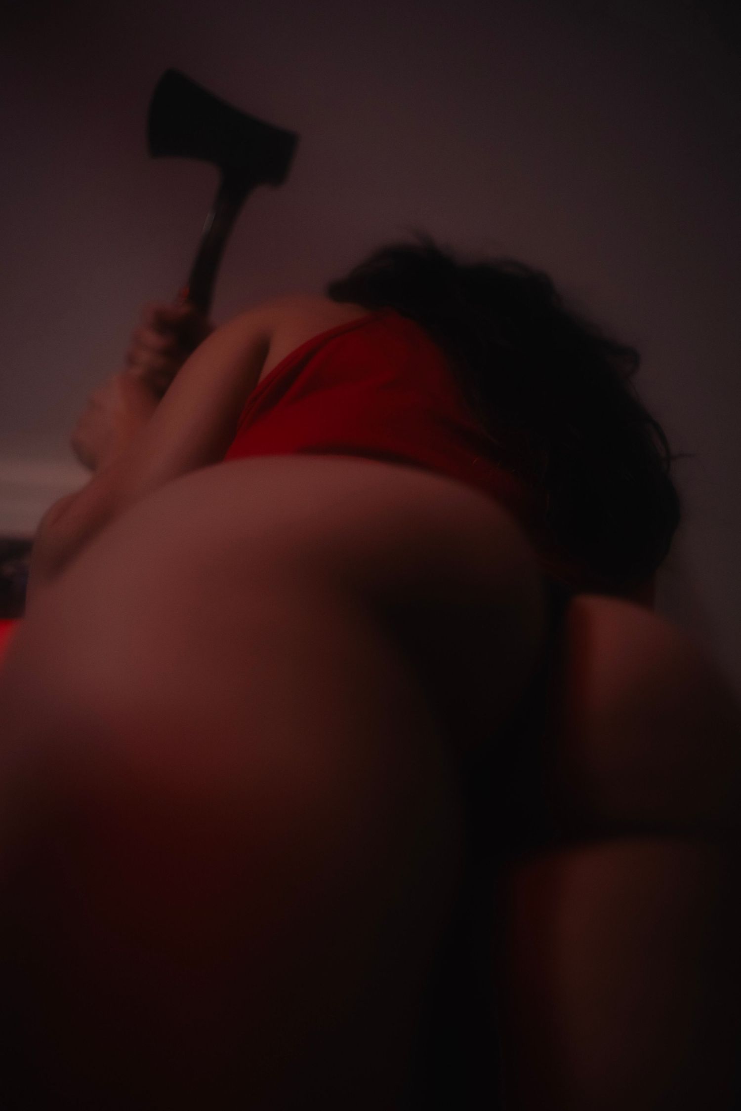 A person in red lingerie holds an axe in a dark, moody low-light artistic horror-themed photo.